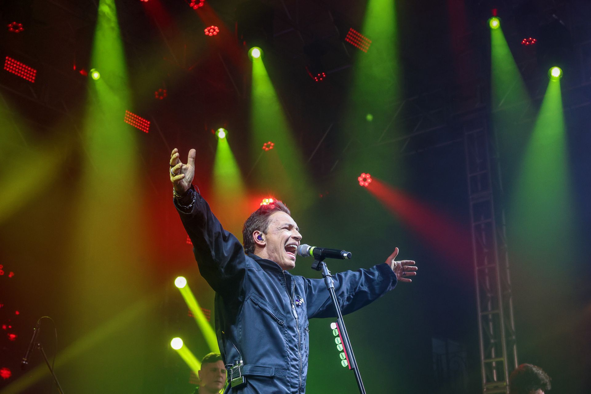 Um cantor com os braços erguidos se apresenta em um palco iluminado por holofotes verdes e vermelhos brilhantes.