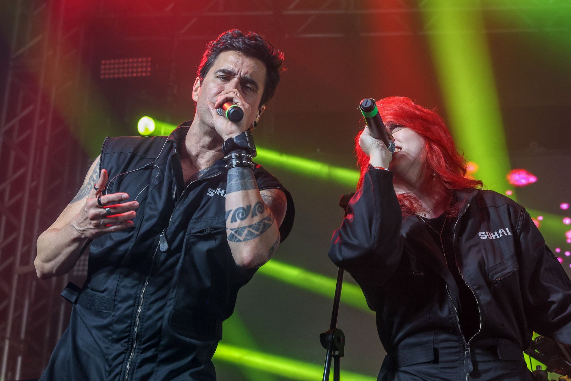 Dois artistas vestidas com macacões pretos cantam em microfones sob luzes de palco verde-brilhante.
