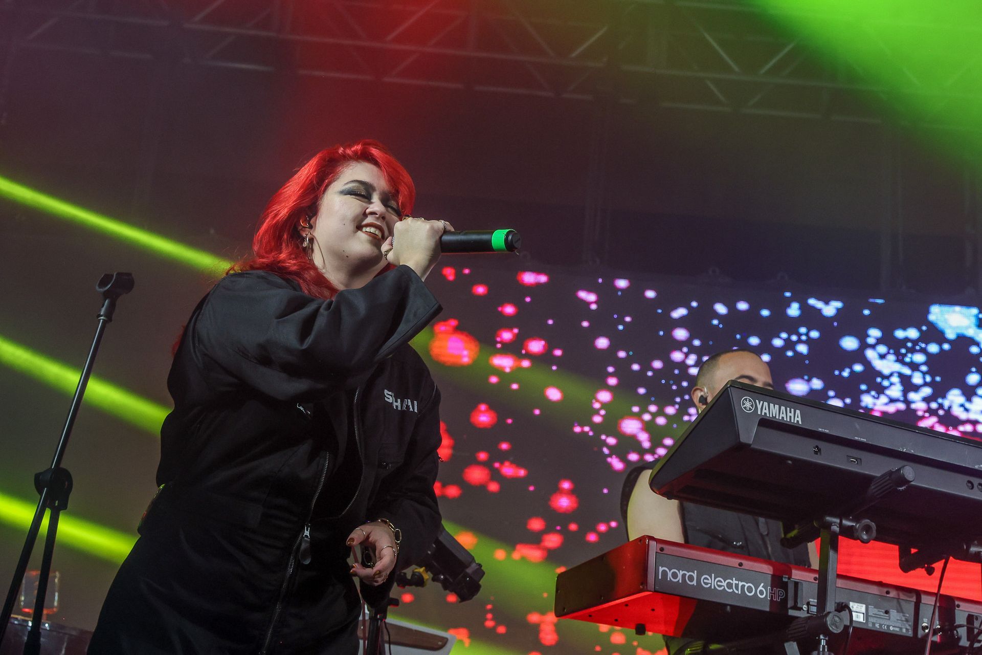 Uma artista de cabelos ruivos vibrantes cantando em um microfone no palco, com um tecladista visível ao fundo.