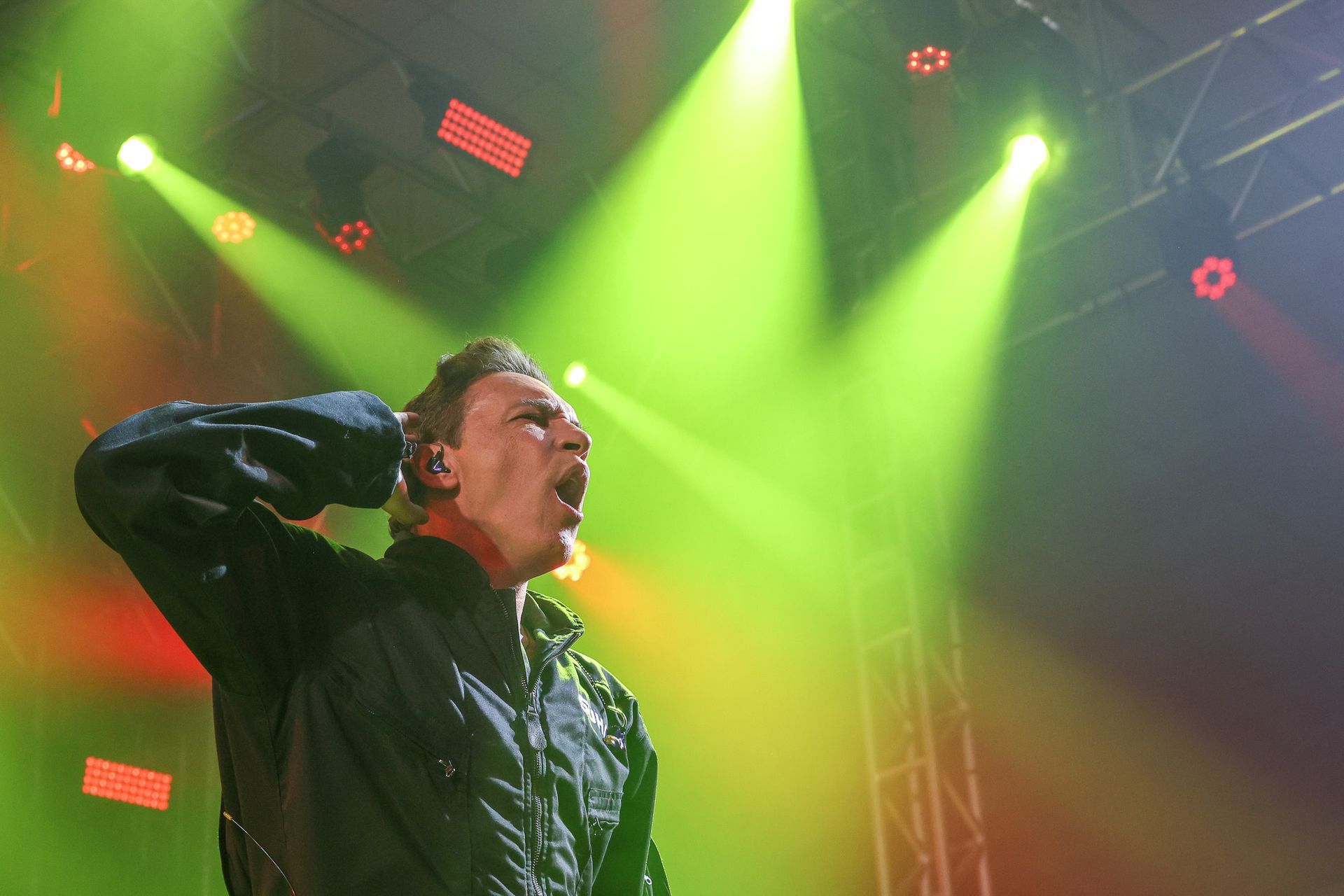 Um cantor se apresenta intensamente no palco sob holofotes amarelos brilhantes, gesticulando em direção à própria orelha.