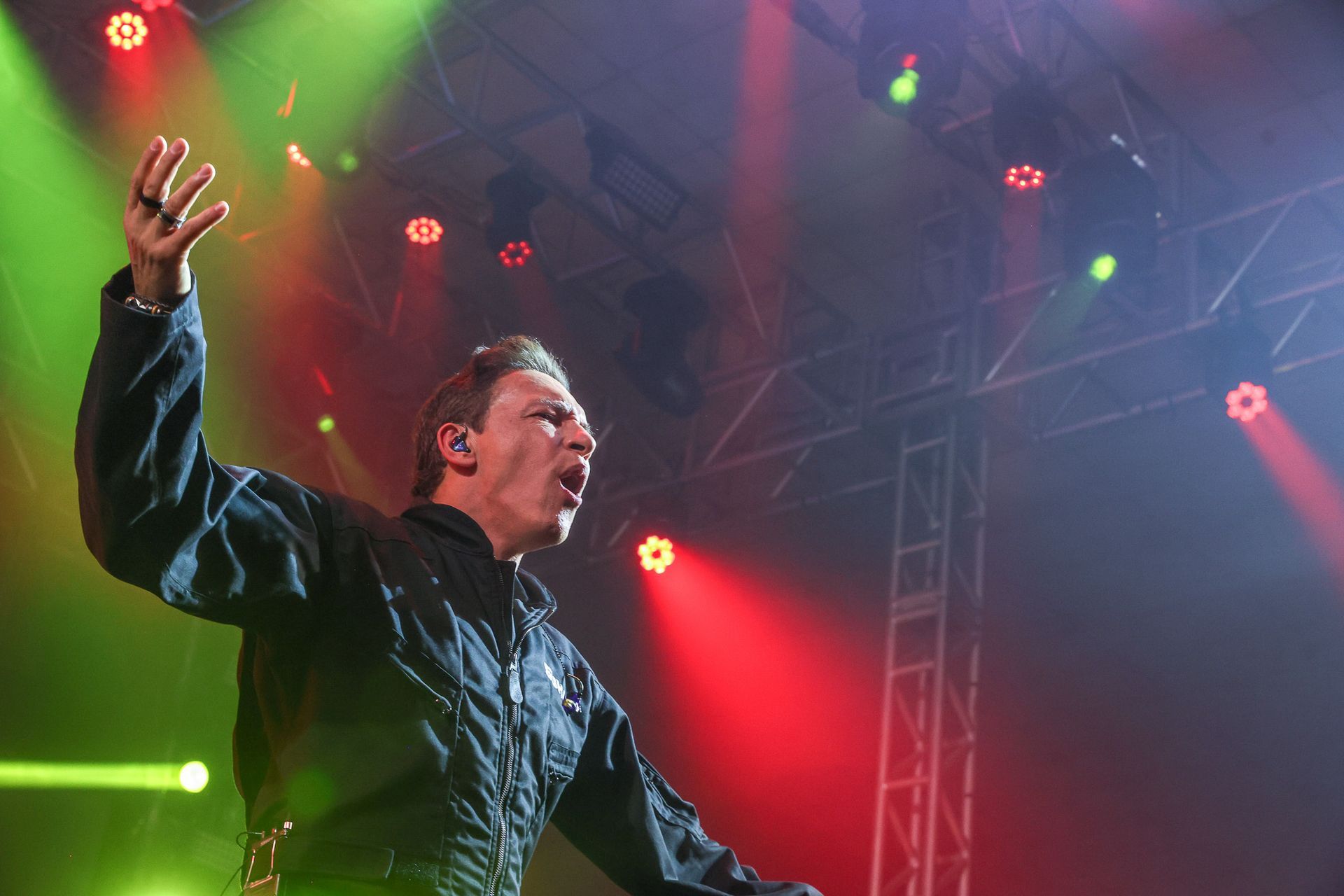 Um artista cantando com o braço levantado em um palco iluminado por holofotes vermelhos e verdes.