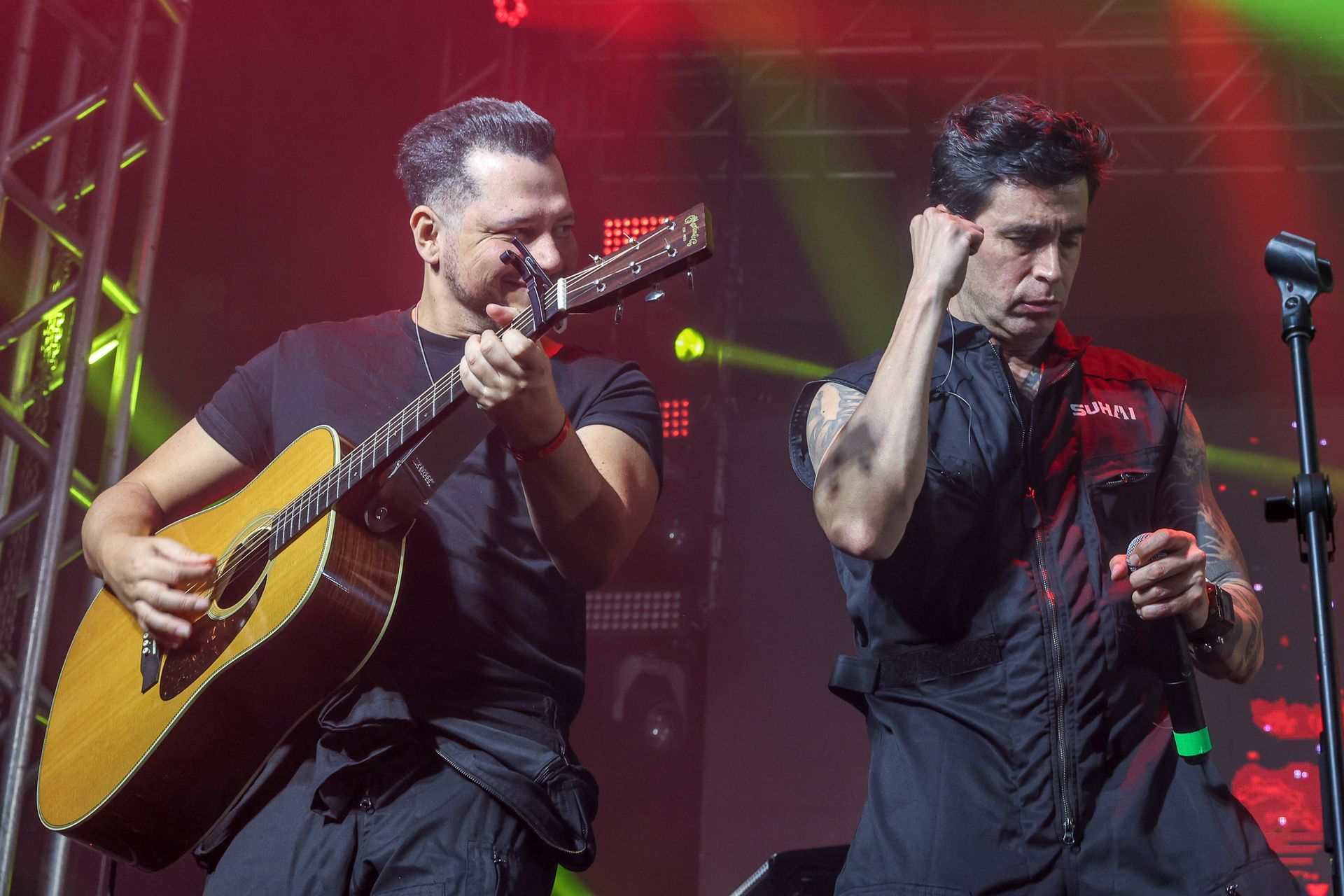 Dois músicos no palco: um toca violão enquanto o outro está em pé com um microfone, ambos vestidos de preto.