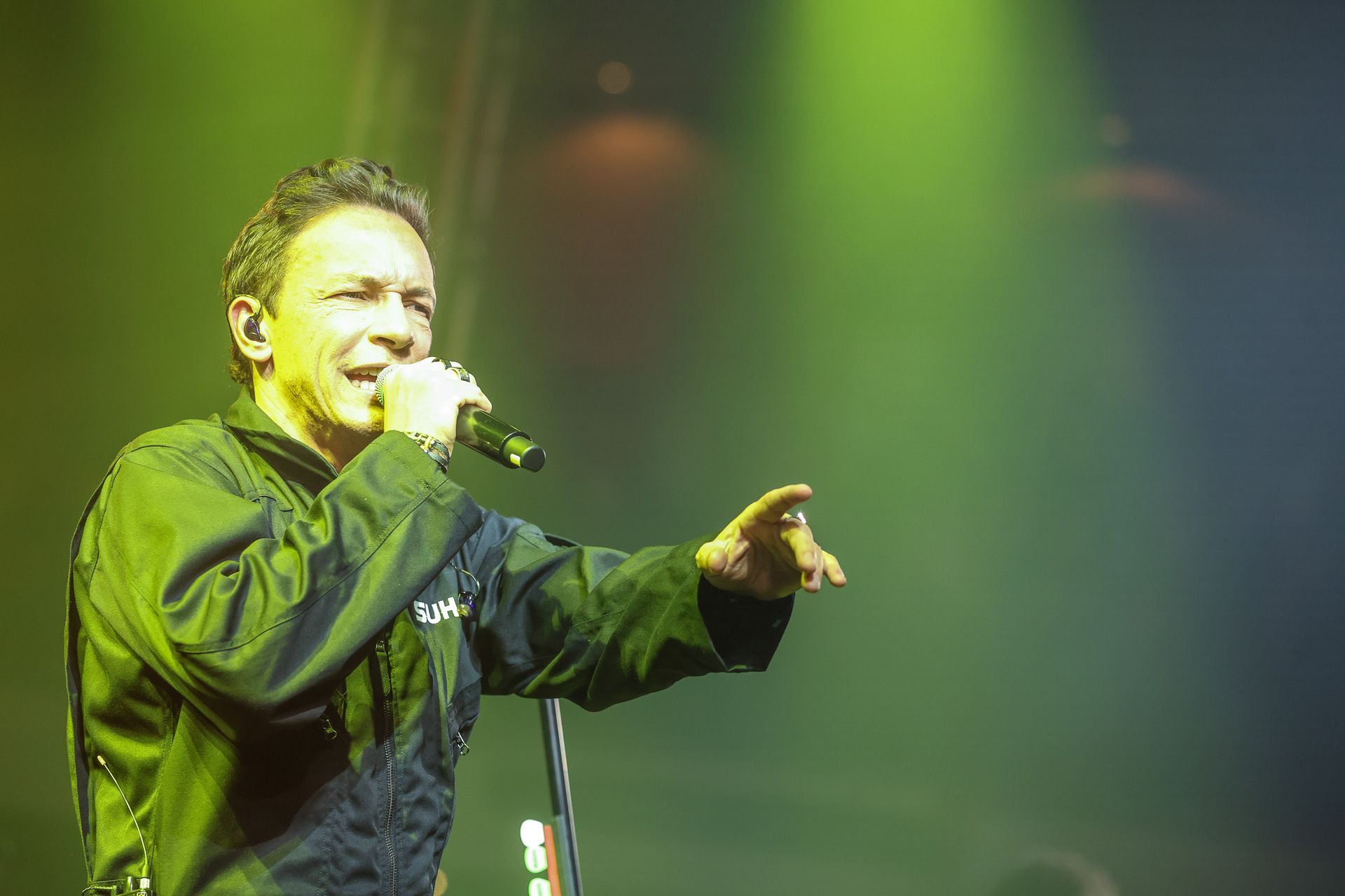 Um artista vestindo um casaco escuro canta em um microfone no palco, sob uma iluminação verde brilhante.
