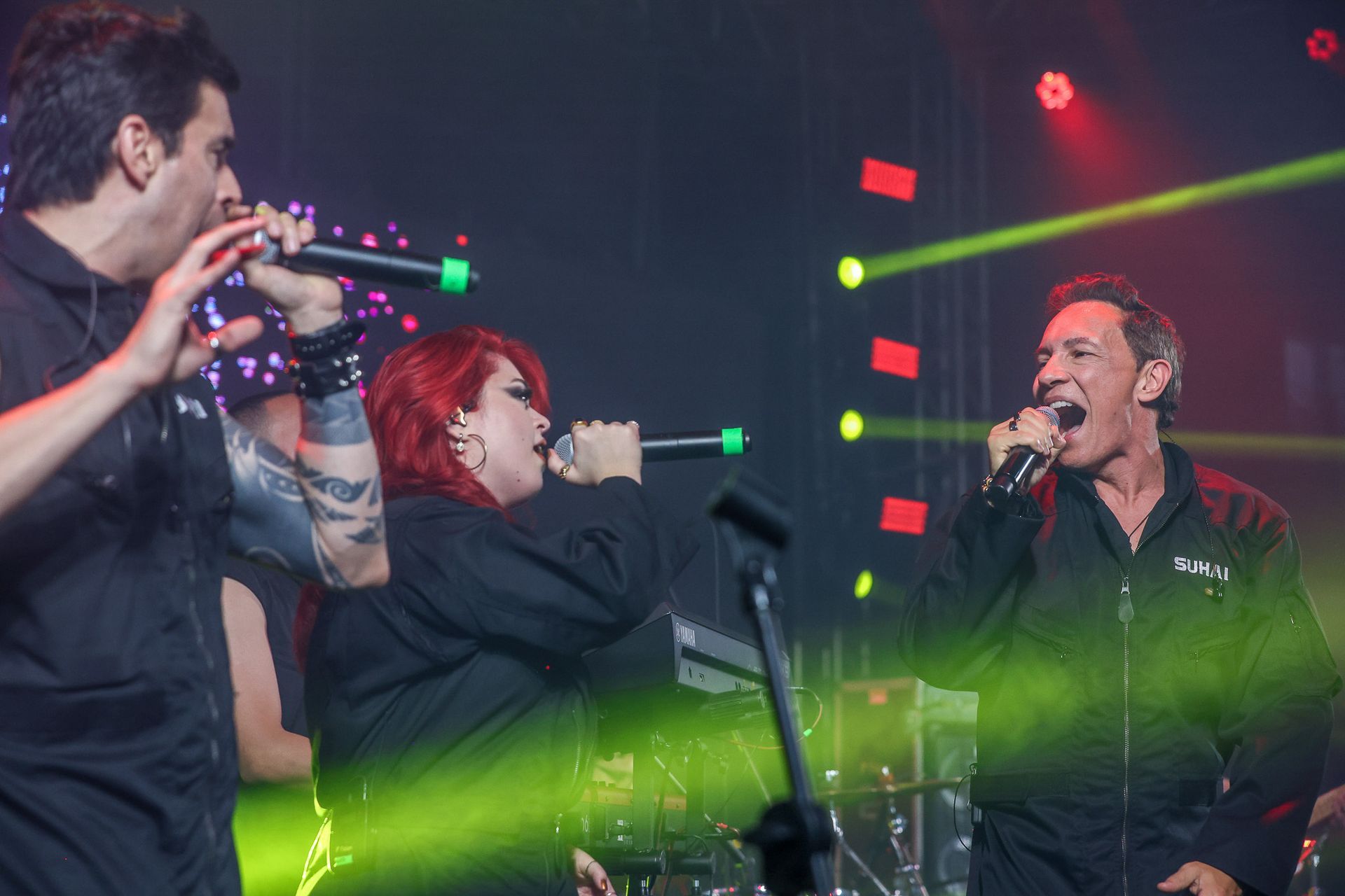 Três pessoas se apresentam em um palco de shows, cantando em microfones sob luzes de palco brilhantes e lasers.