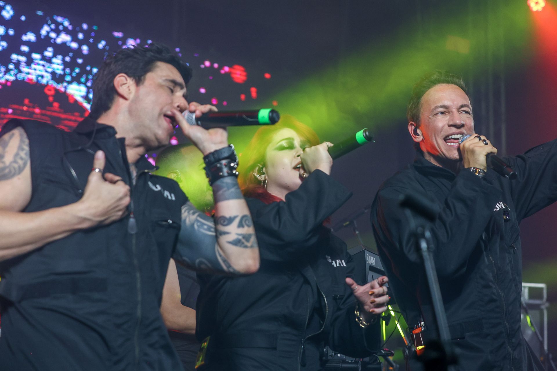 Três artistas com uniformes pretos cantam em microfones num palco com iluminação vibrante em tons de verde e vermelho.