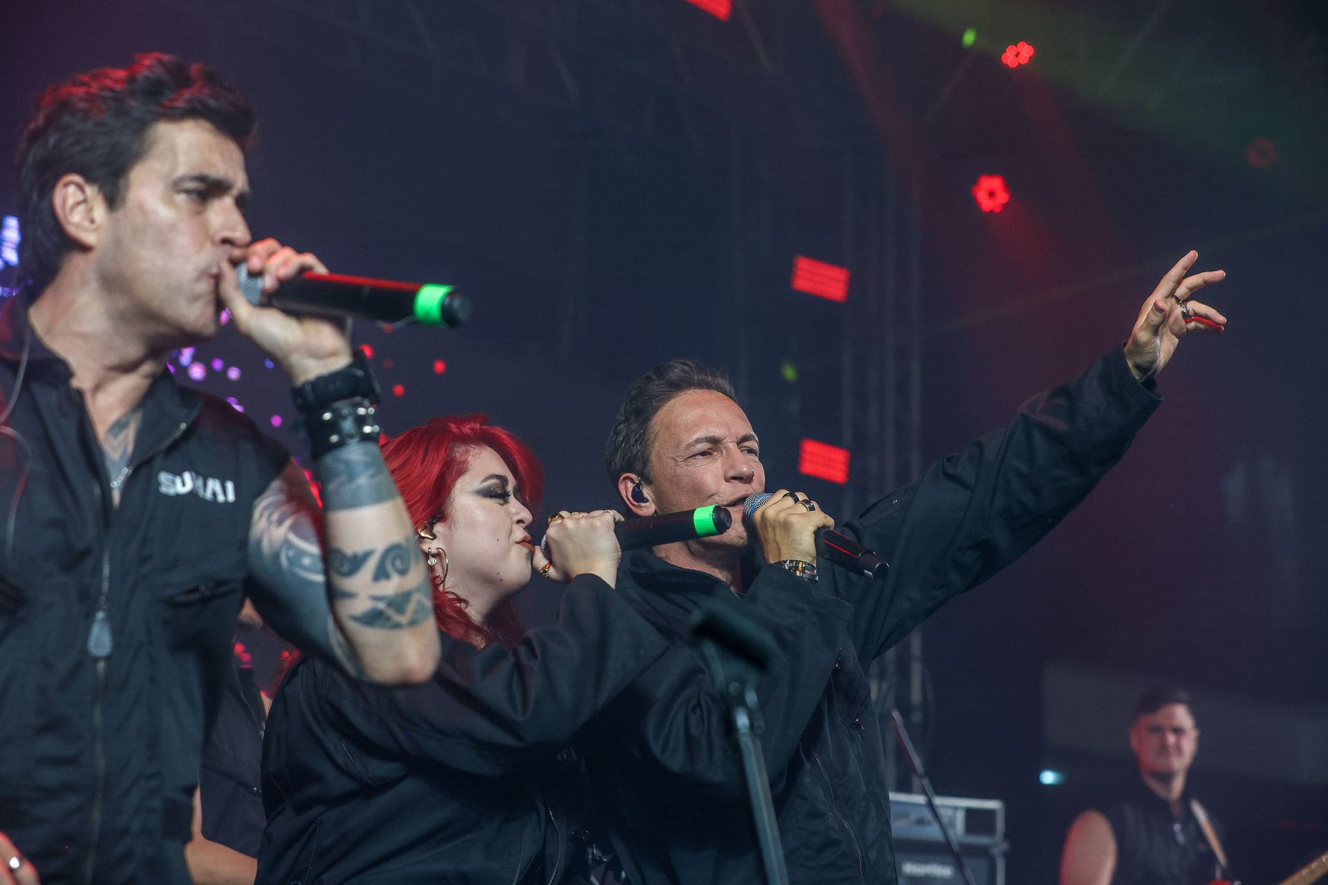 Três artistas vestidos com trajes de palco pretos cantam em microfones sob luzes vermelhas de concerto.