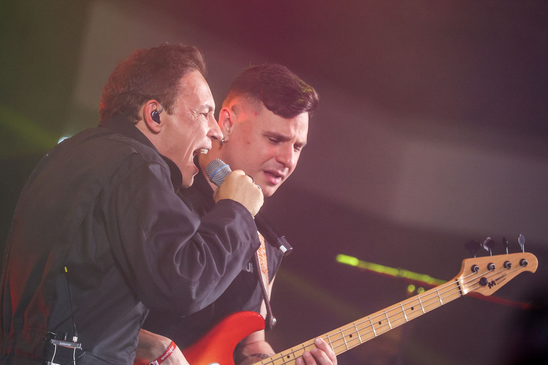 Dois artistas no palco: um cantando intensamente em um microfone enquanto o outro toca um baixo elétrico vermelho.