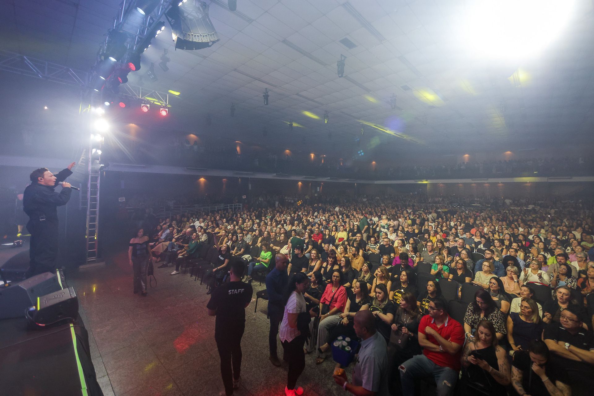 Um artista está em um palco dirigindo-se a uma grande plateia sentada em um auditório com pouca luz, sob fortes holofotes.