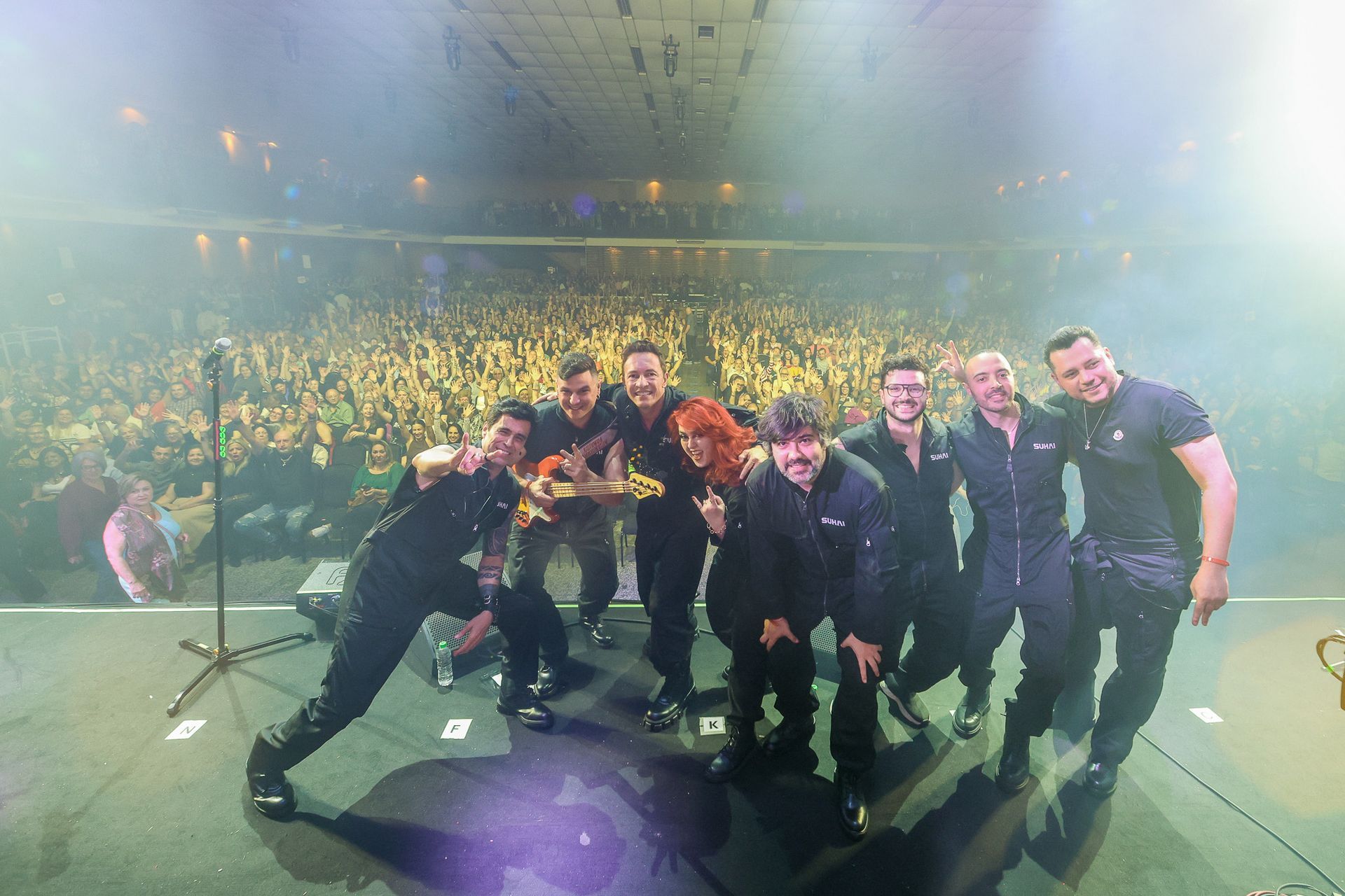 Uma banda com uniformes pretos posa em um palco diante de uma grande plateia, iluminada por luzes de palco brilhantes.