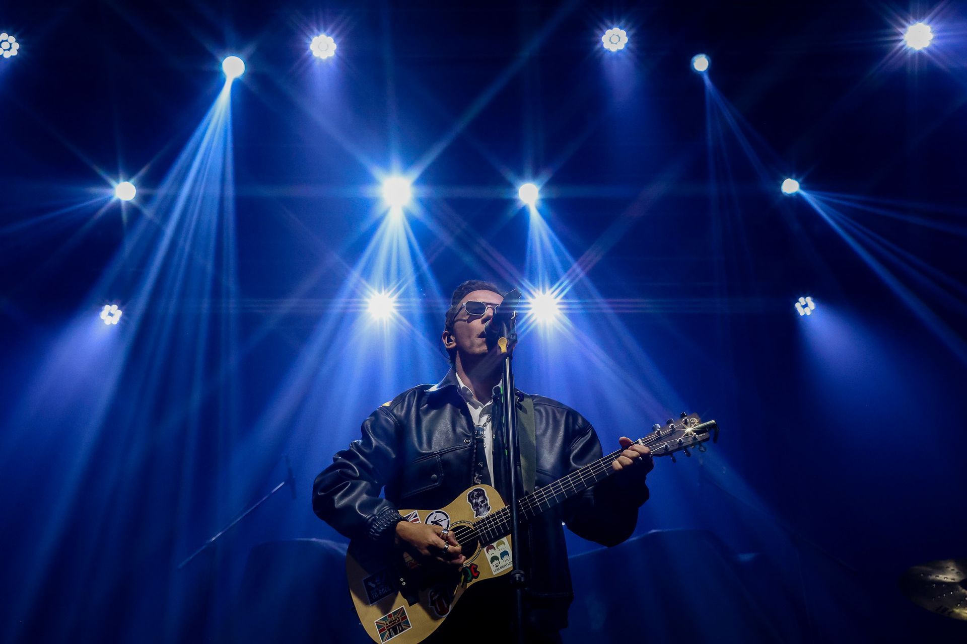 Uma pessoa vestindo uma jaqueta de couro toca violão enquanto canta em um microfone em um palco com luzes azuis brilhantes.