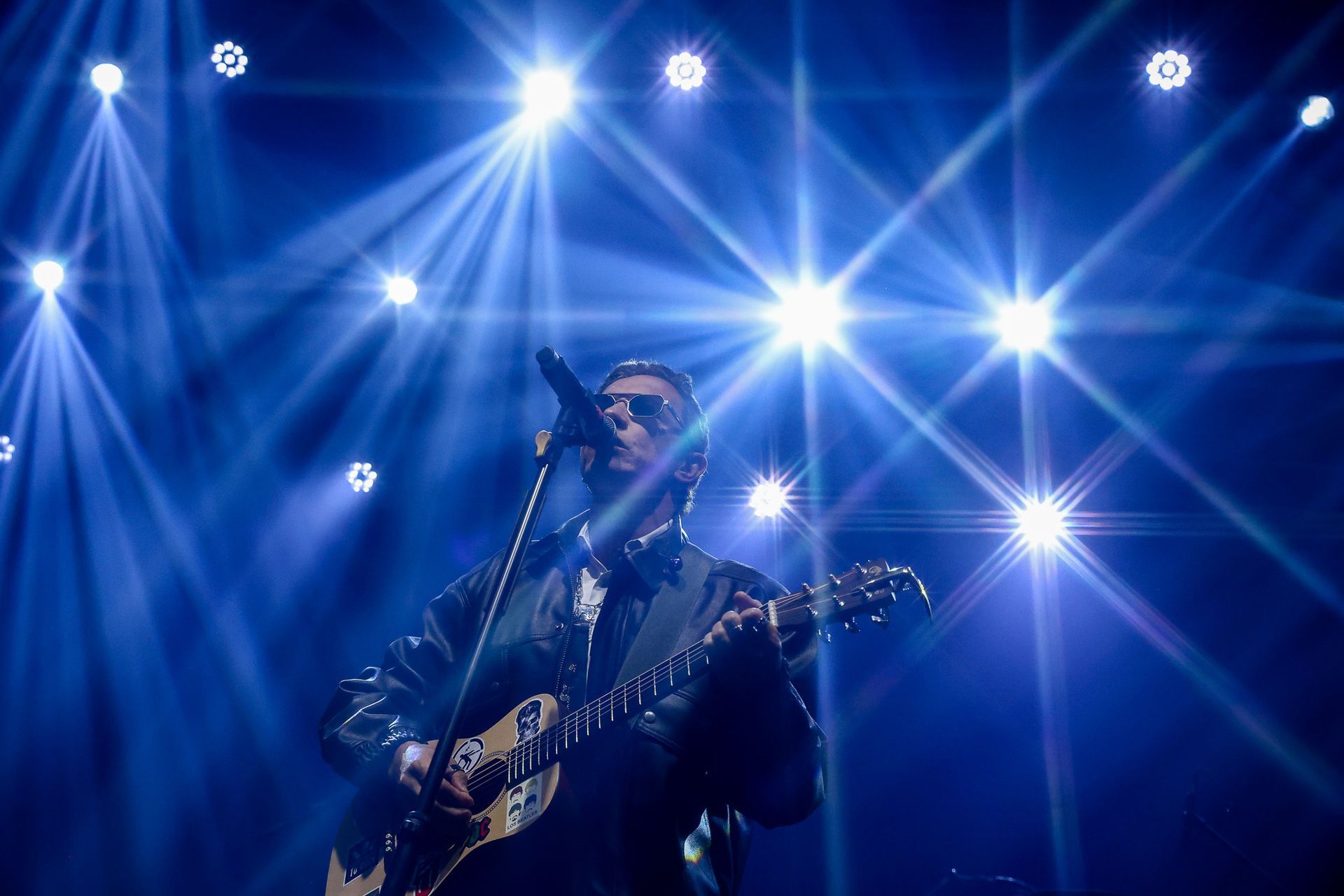 Um artista tocando violão e cantando em um microfone em um palco iluminado por luzes azuis brilhantes com efeito de estrela.