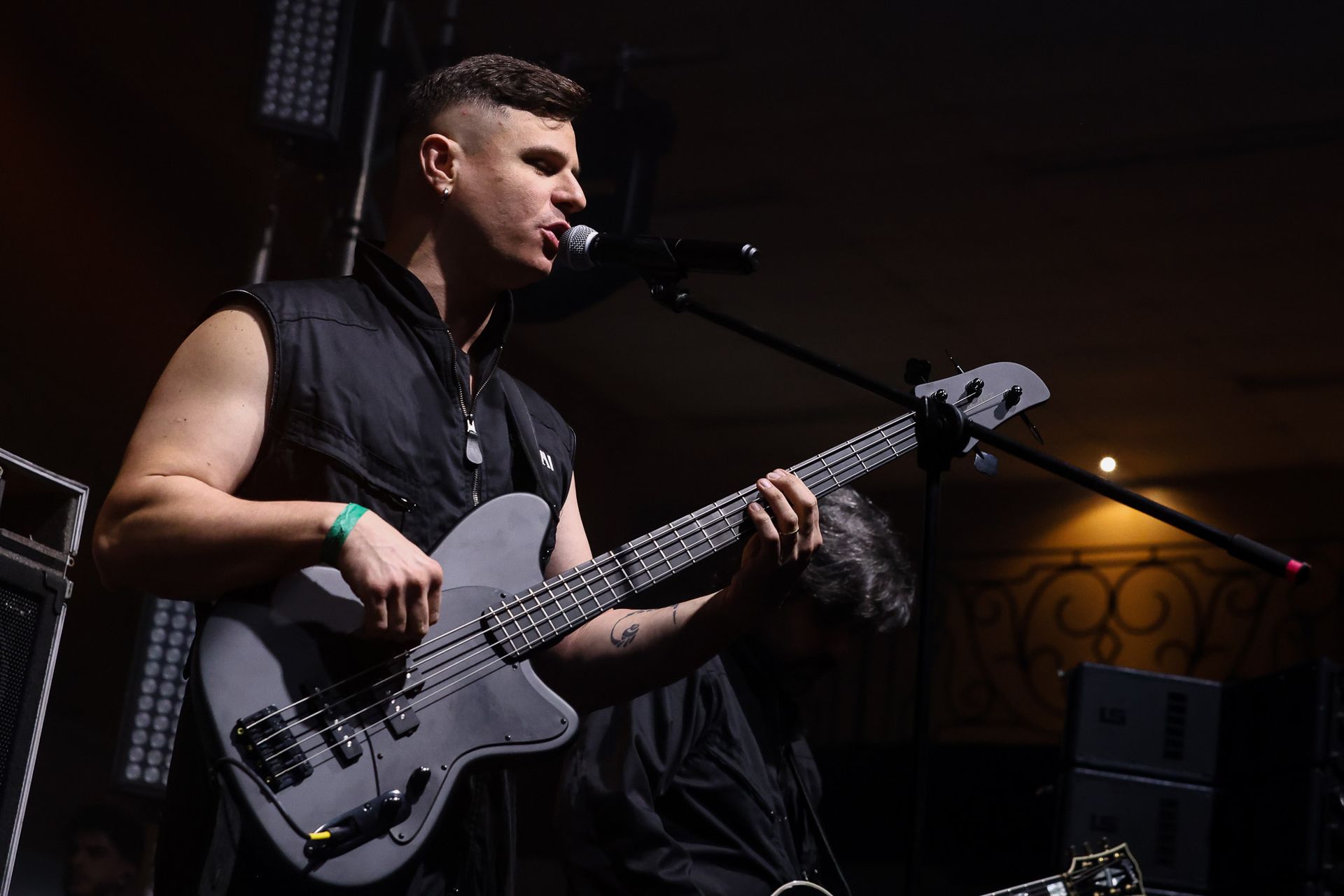Um músico de cabelo curto se apresenta no palco, tocando um baixo cinza enquanto canta ao microfone.