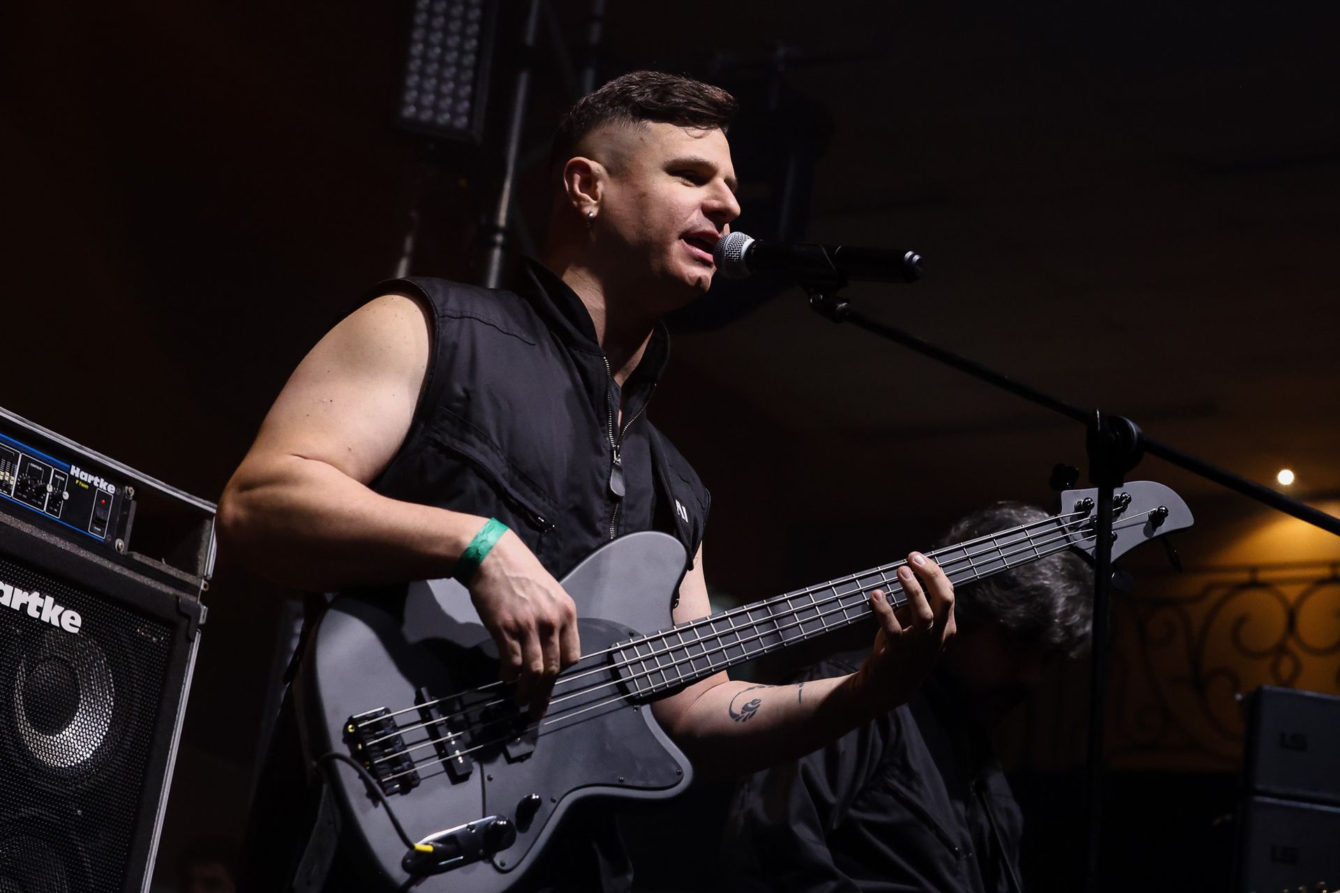 Um músico vestindo um colete preto sem mangas toca um baixo cinza enquanto canta em um microfone em um palco escuro.