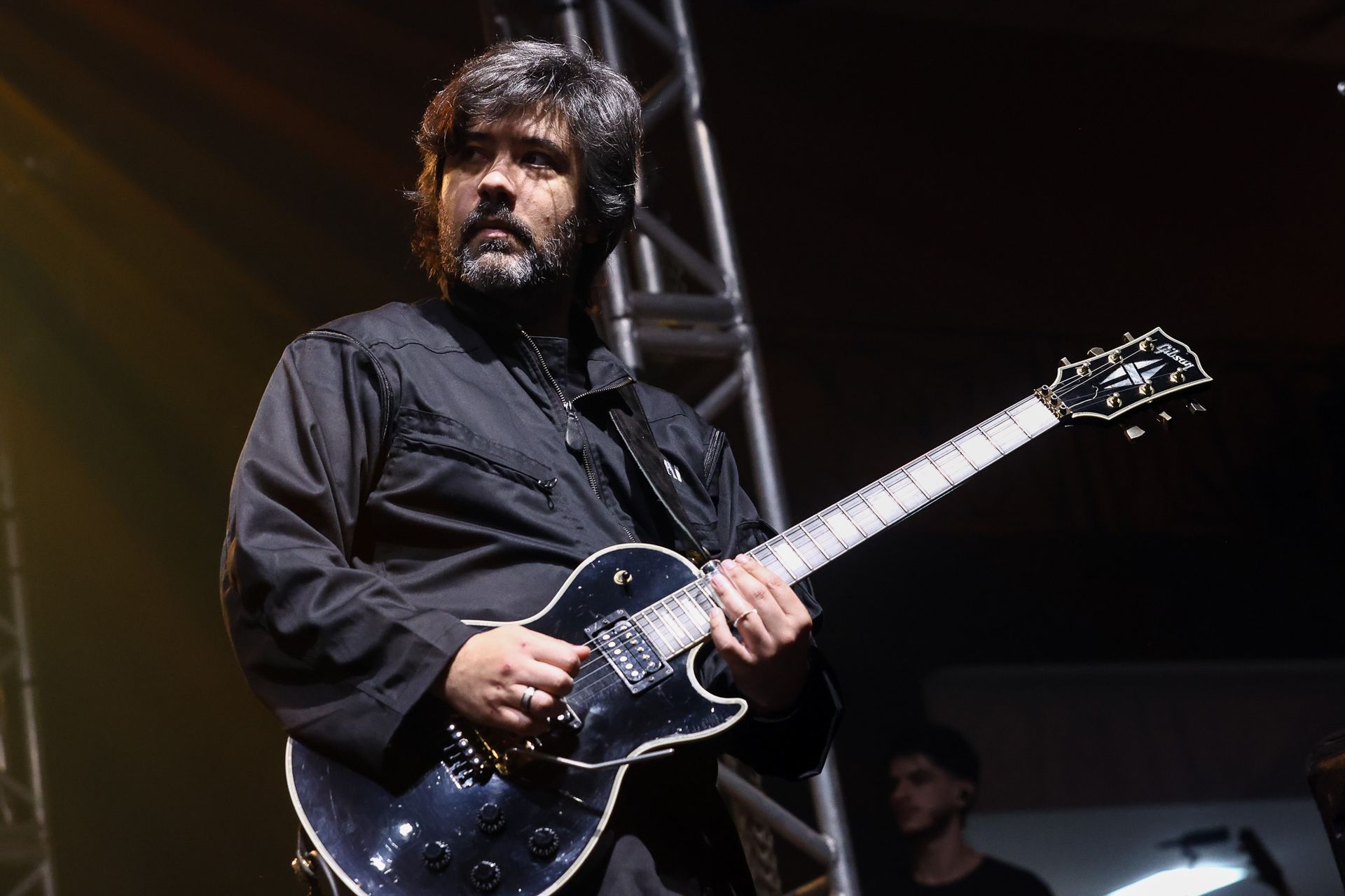 Um músico barbudo, vestindo uma camisa preta, toca uma guitarra elétrica em um palco com uma estrutura metálica ao fundo.