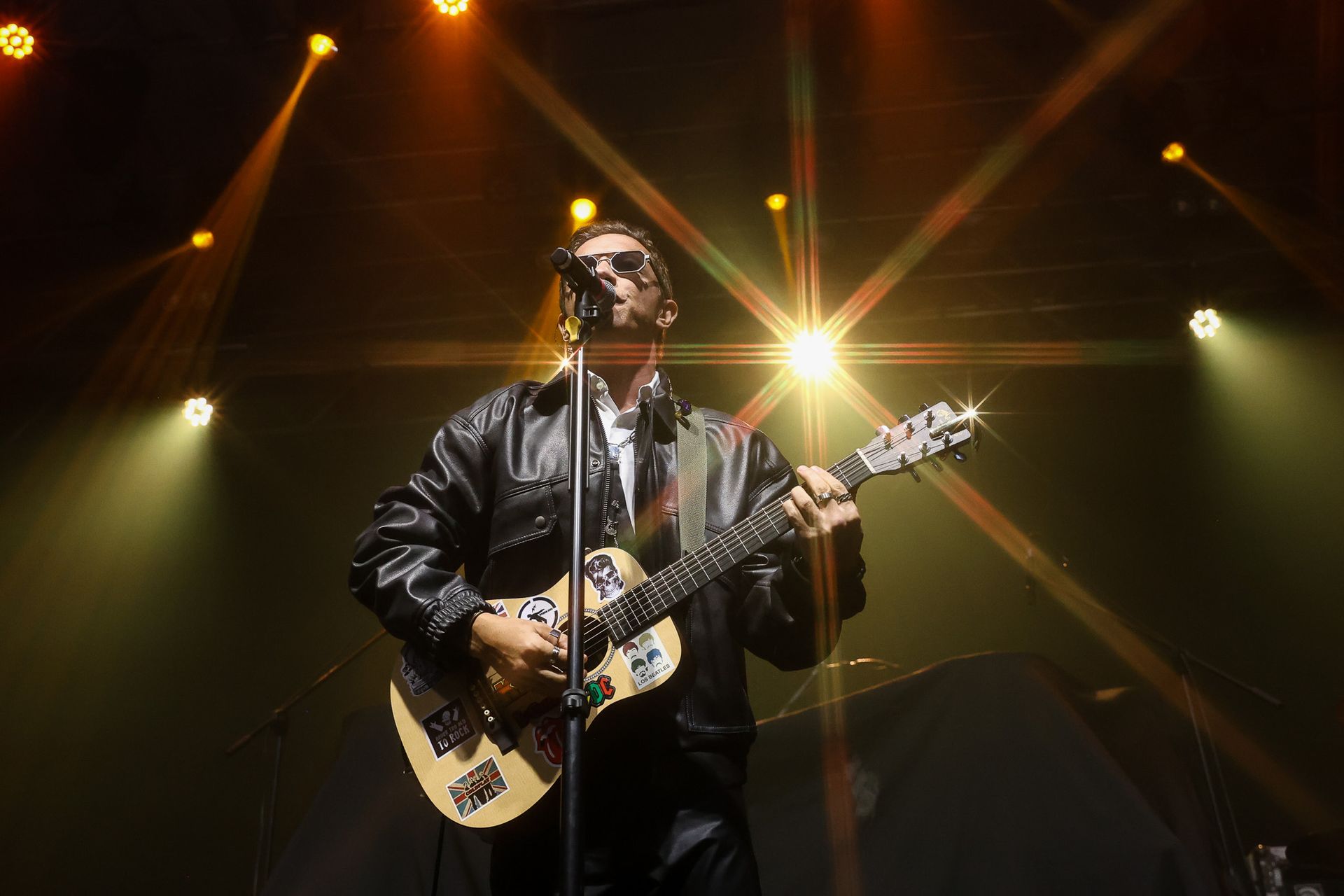 Um artista vestindo uma jaqueta de couro preta toca violão em um microfone em um palco iluminado por luzes brilhantes 