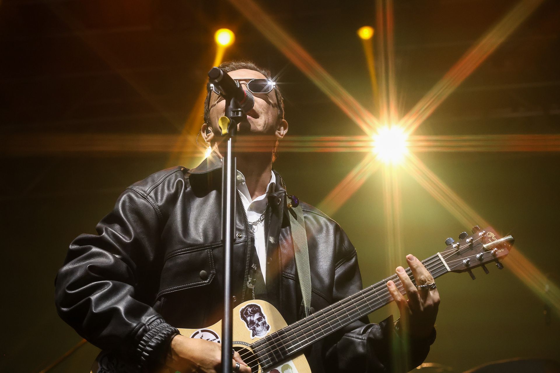 Um músico vestindo jaqueta de couro preta e óculos escuros canta em um microfone enquanto toca violão no palco.