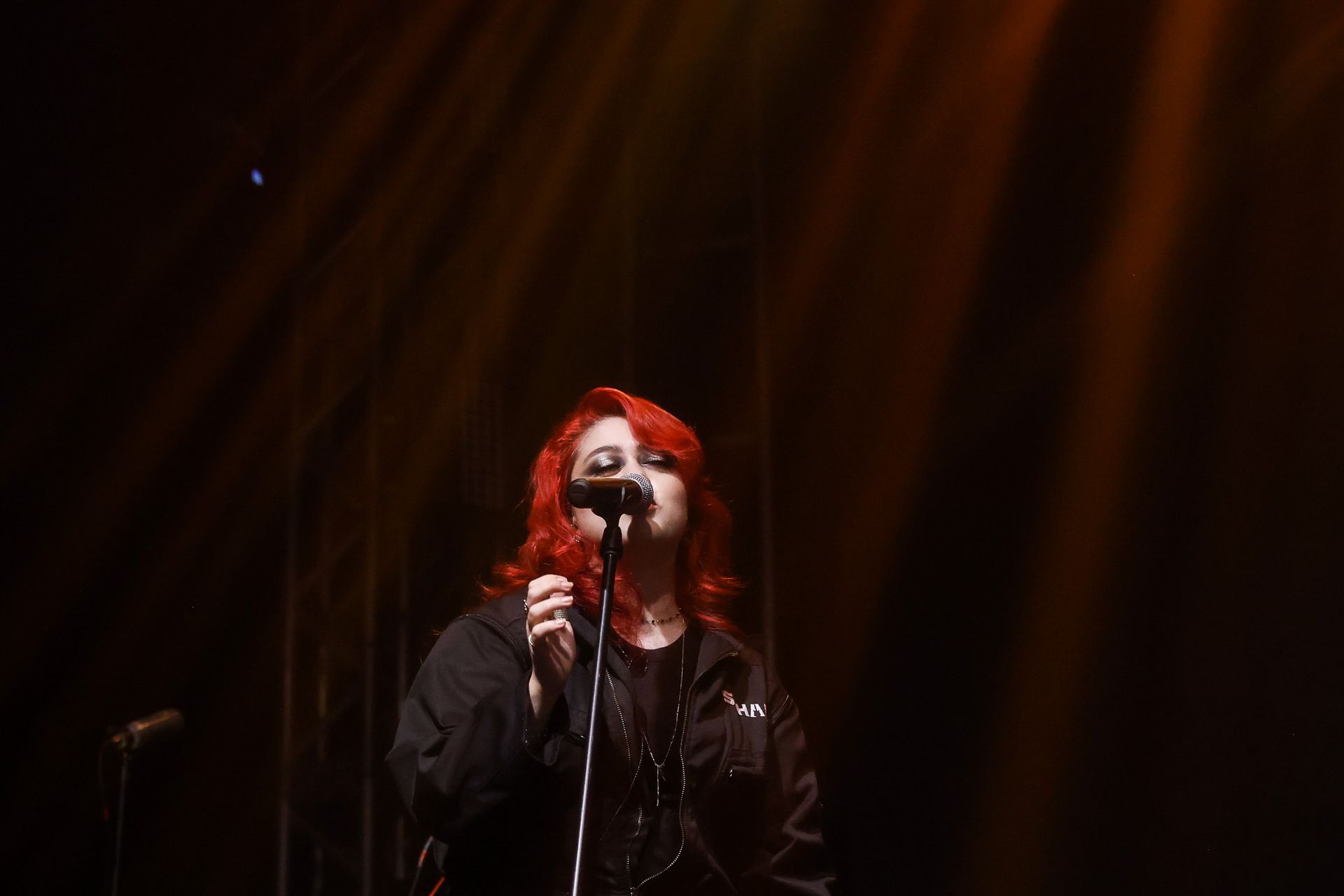 Uma artista de cabelos ruivos cantando em um microfone em um palco pouco iluminado, sob holofotes dramáticos de tons quentes.