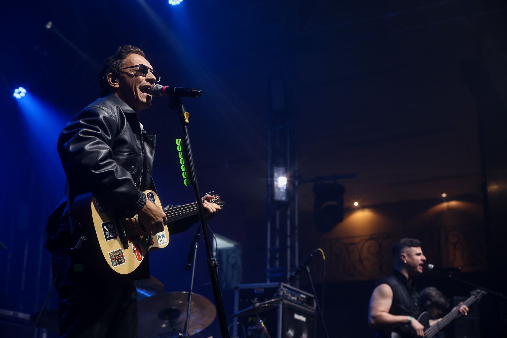 Um músico cantando ao microfone e tocando violão em um palco de concerto com pouca luz, com um baixista ao fundo.