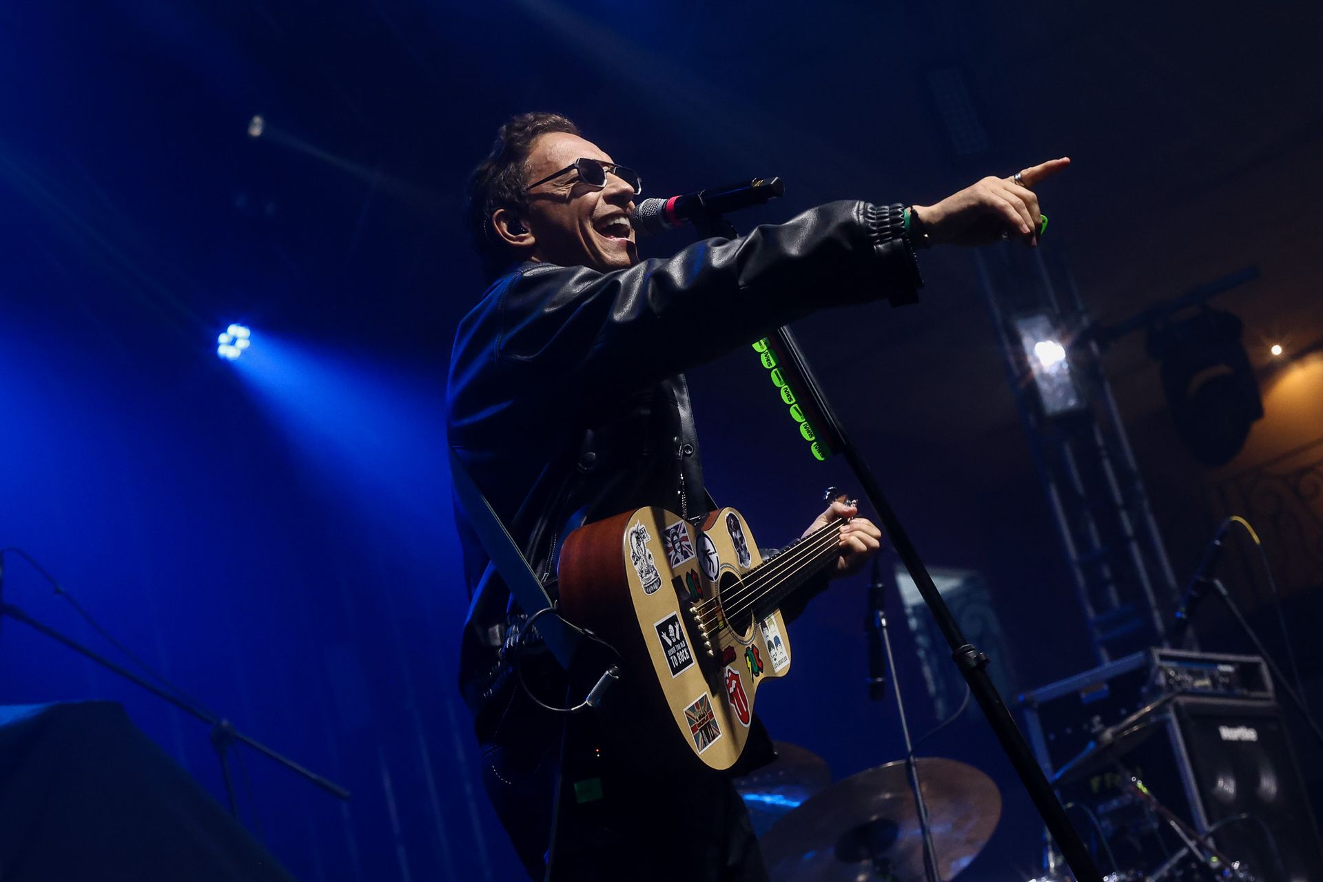 Um artista em um palco escuro, vestindo uma jaqueta de couro, toca um violão coberto de adesivos enquanto aponta para o lado.