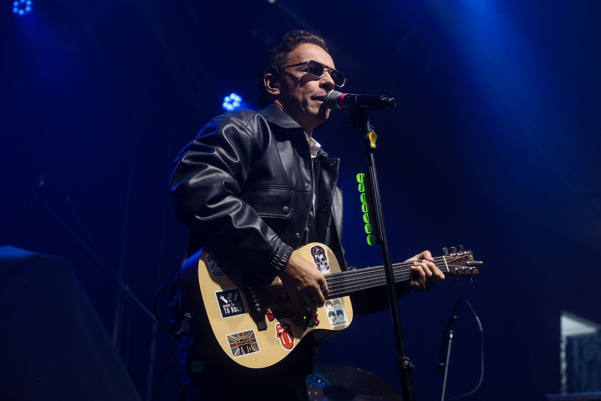 Um músico vestindo jaqueta de couro e óculos escuros toca violão com adesivos enquanto se apresenta no palco.