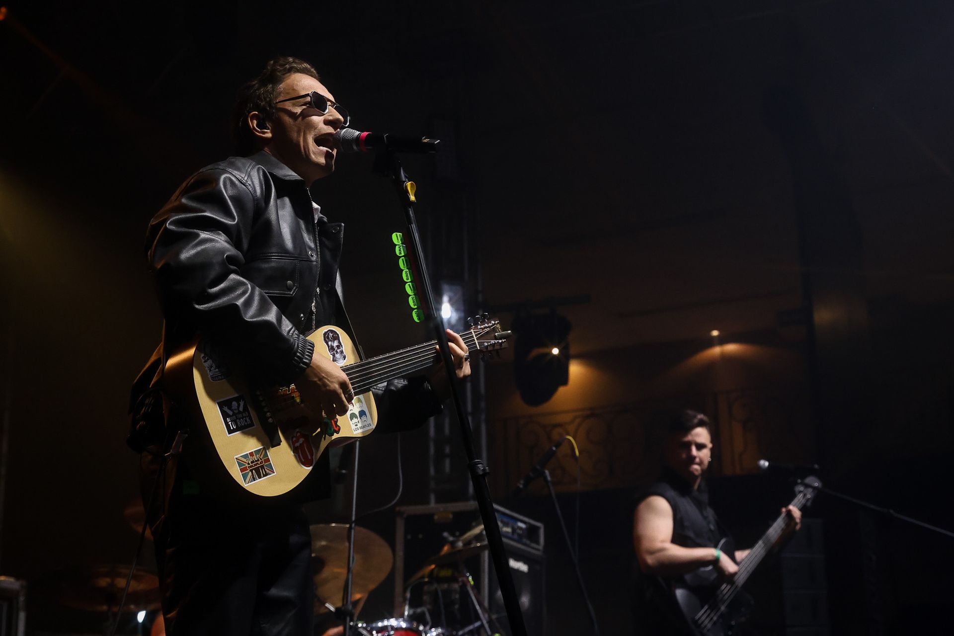 Um músico de jaqueta de couro toca violão e canta ao microfone em um palco de concerto com pouca luz.