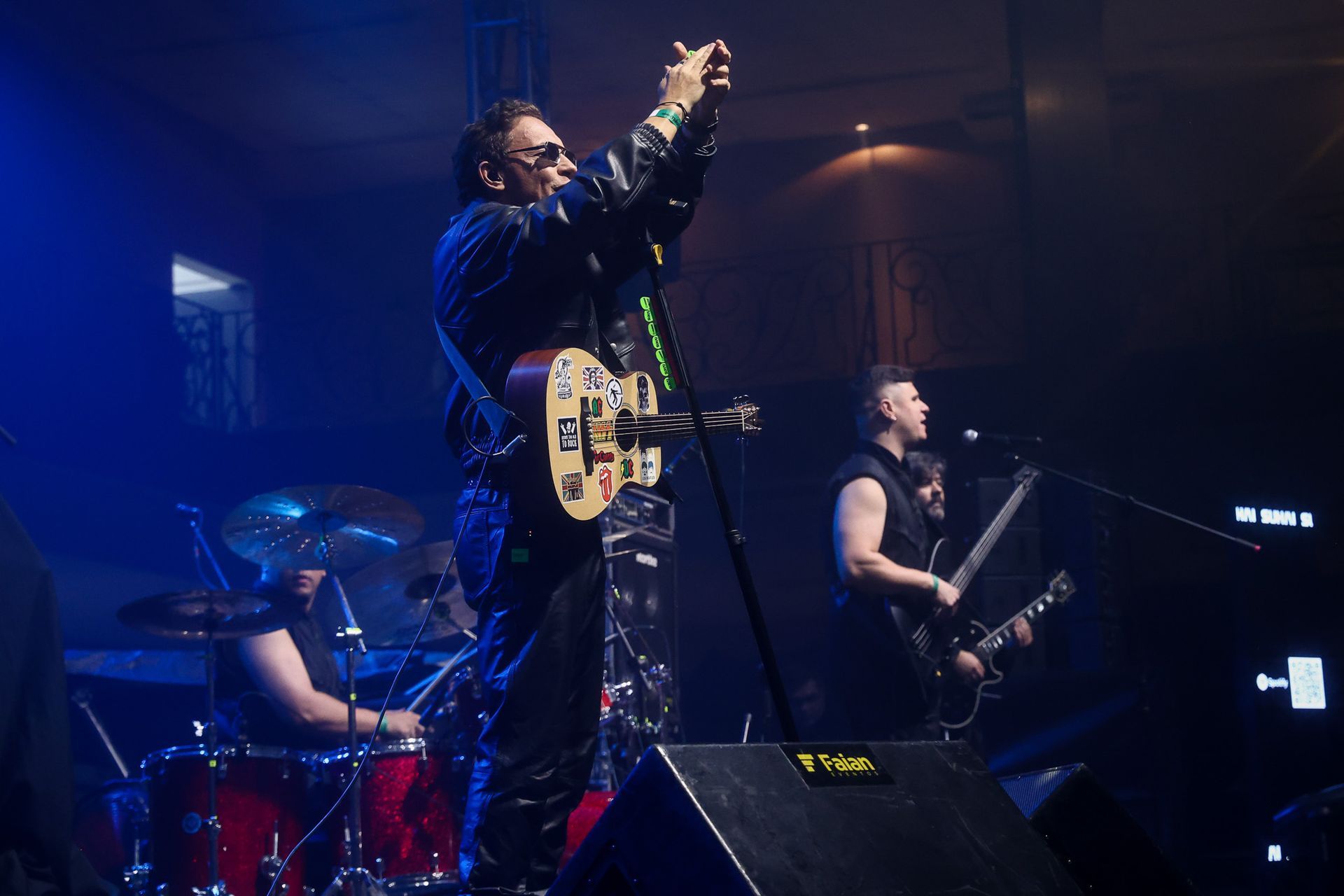Uma banda ao vivo se apresenta no palco sob luzes azuis, enquanto um guitarrista filma a plateia com um celular.