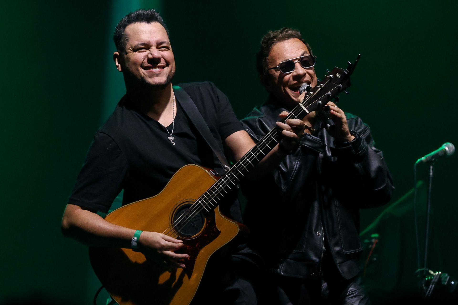 Dois músicos se apresentando no palco, um tocando violão e sorrindo, o outro cantando ao microfone.