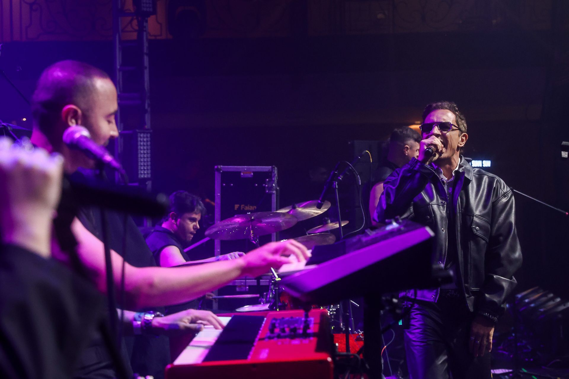 Uma banda se apresenta em um palco com iluminação fraca e luzes roxas, com um vocalista de jaqueta de couro e um tecladista.