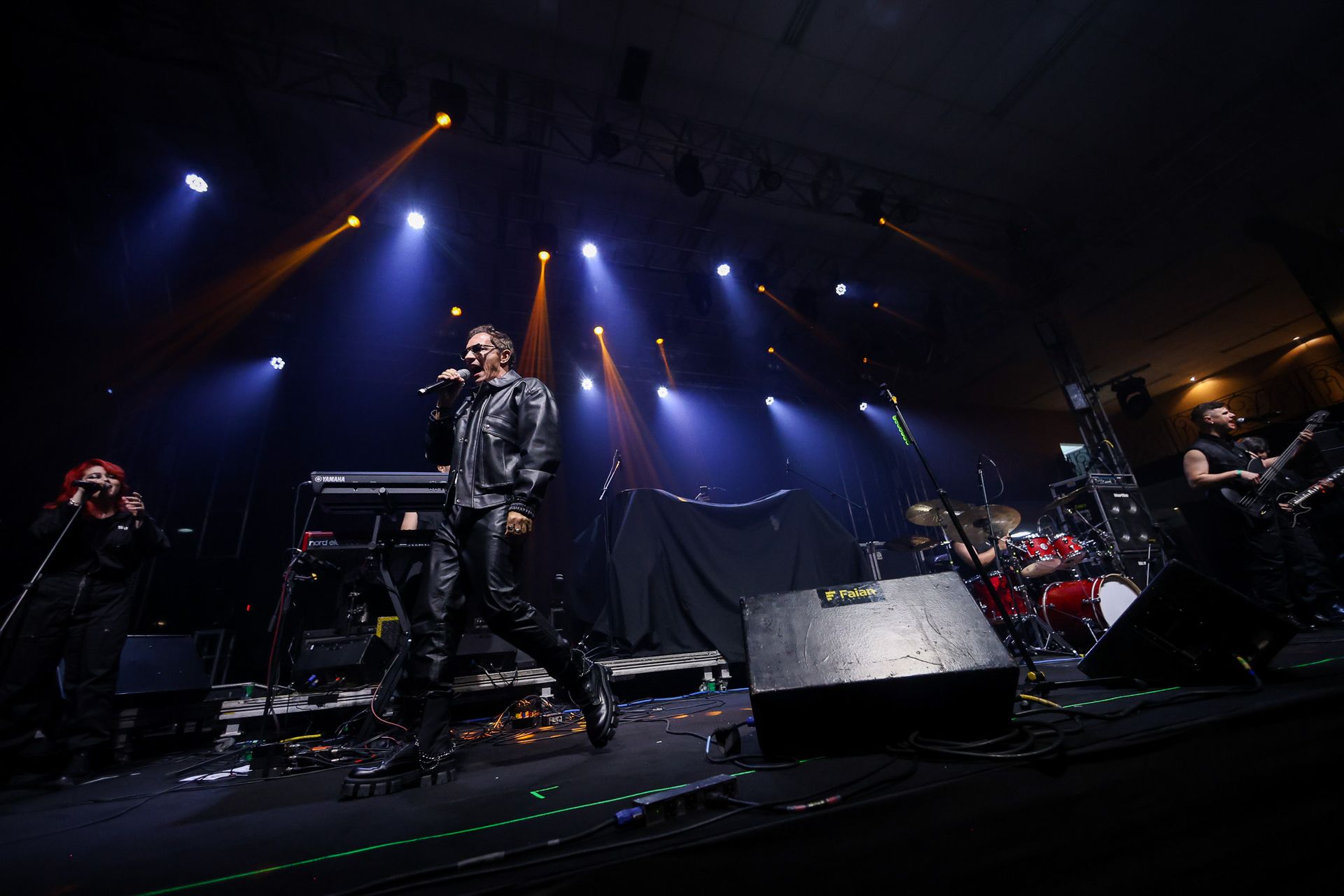 Uma banda, iluminado por holofotes azuis, com um vocalista vestido de couro preto e bateria ao fundo.