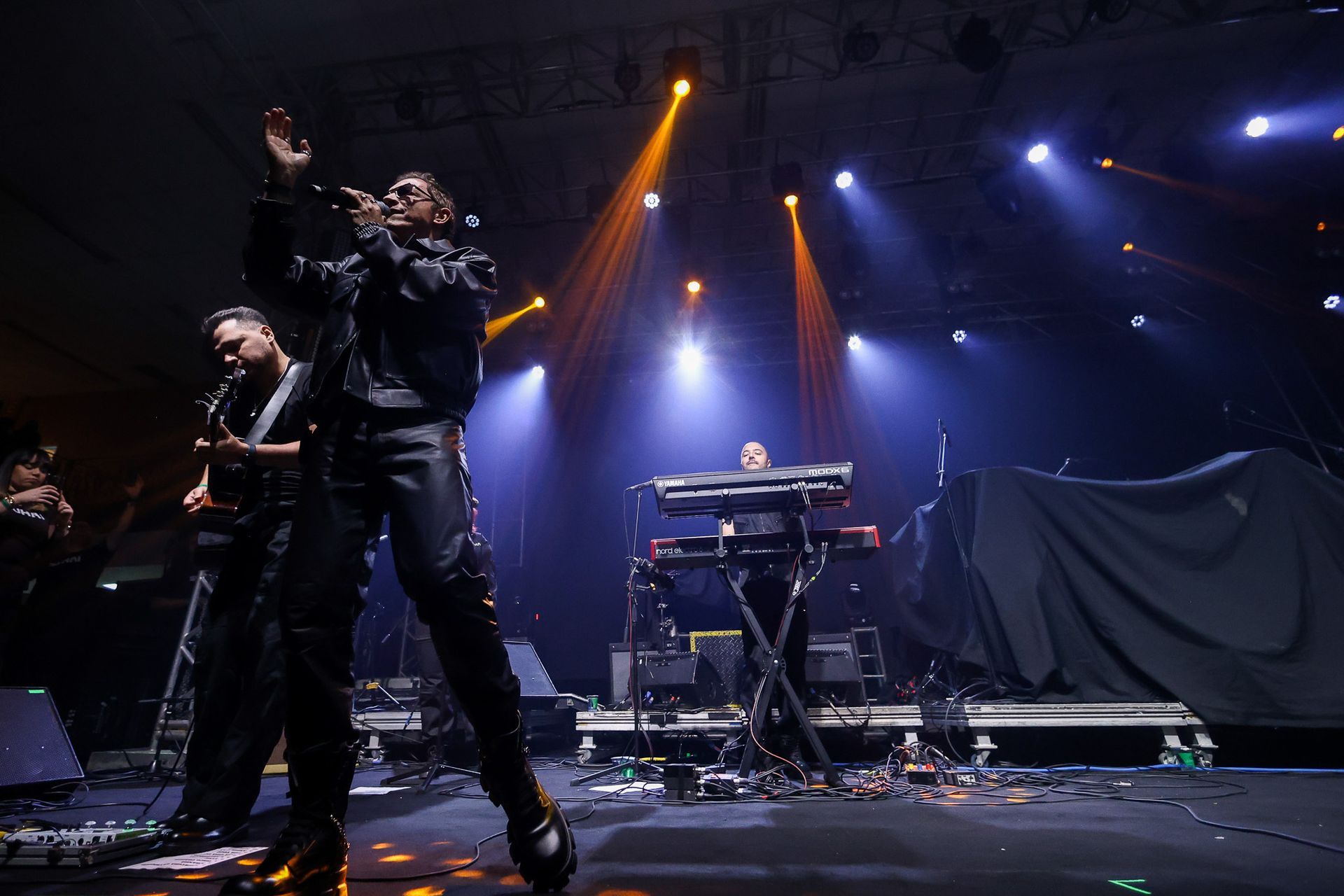 Uma banda se apresenta em um palco com holofotes azuis e laranjas, composta por um vocalista e um tecladista.