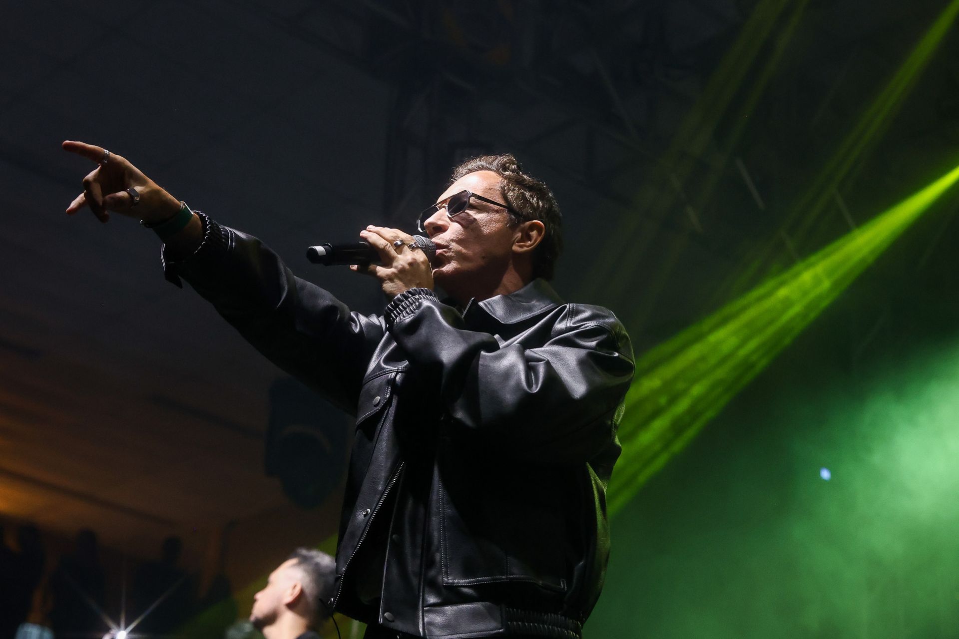 Um artista vestindo uma jaqueta de couro preta canta em um microfone enquanto aponta para o lado.