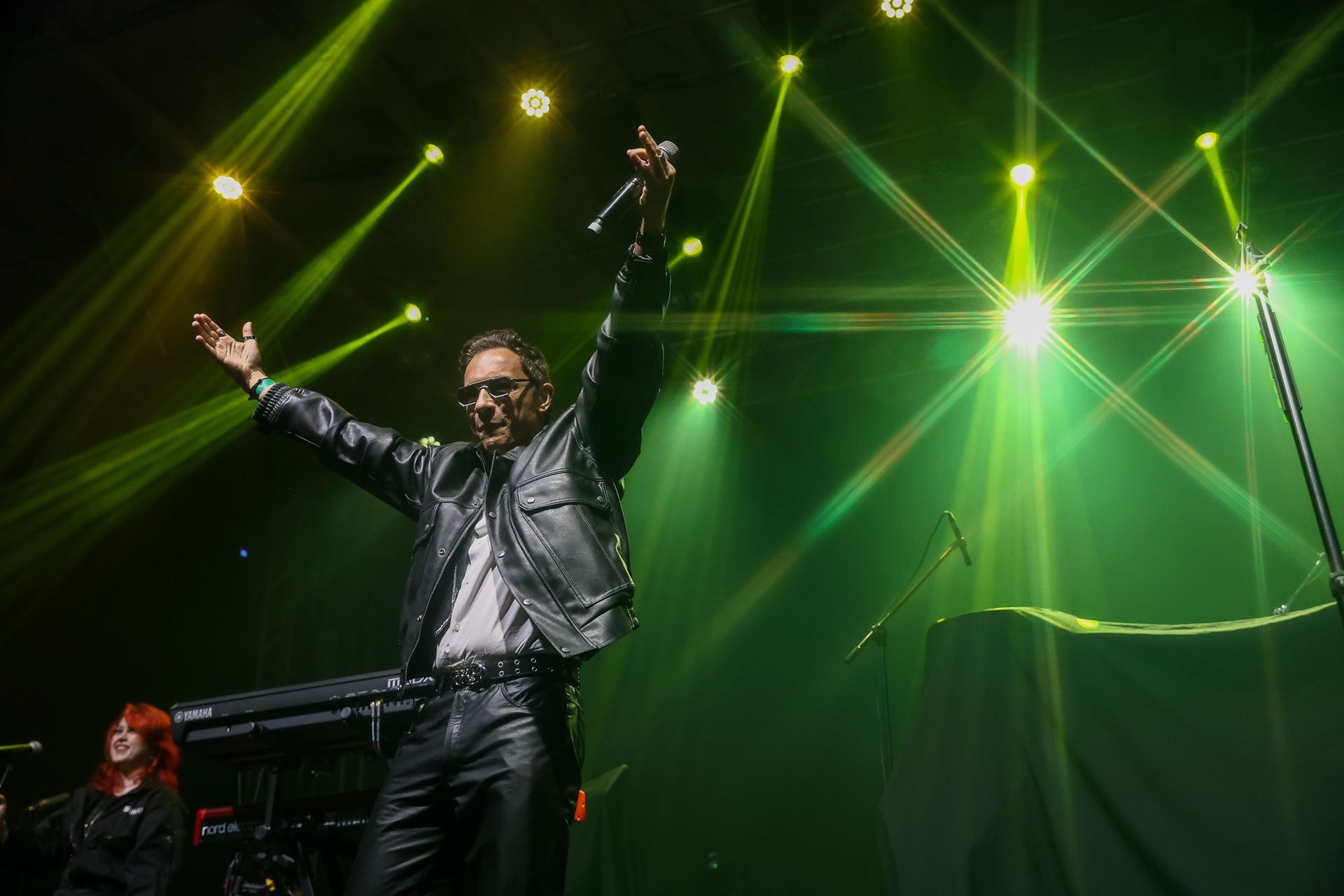 Um artista vestindo jaqueta de couro está no palco com os braços erguidos sob luzes verdes brilhantes de um show.