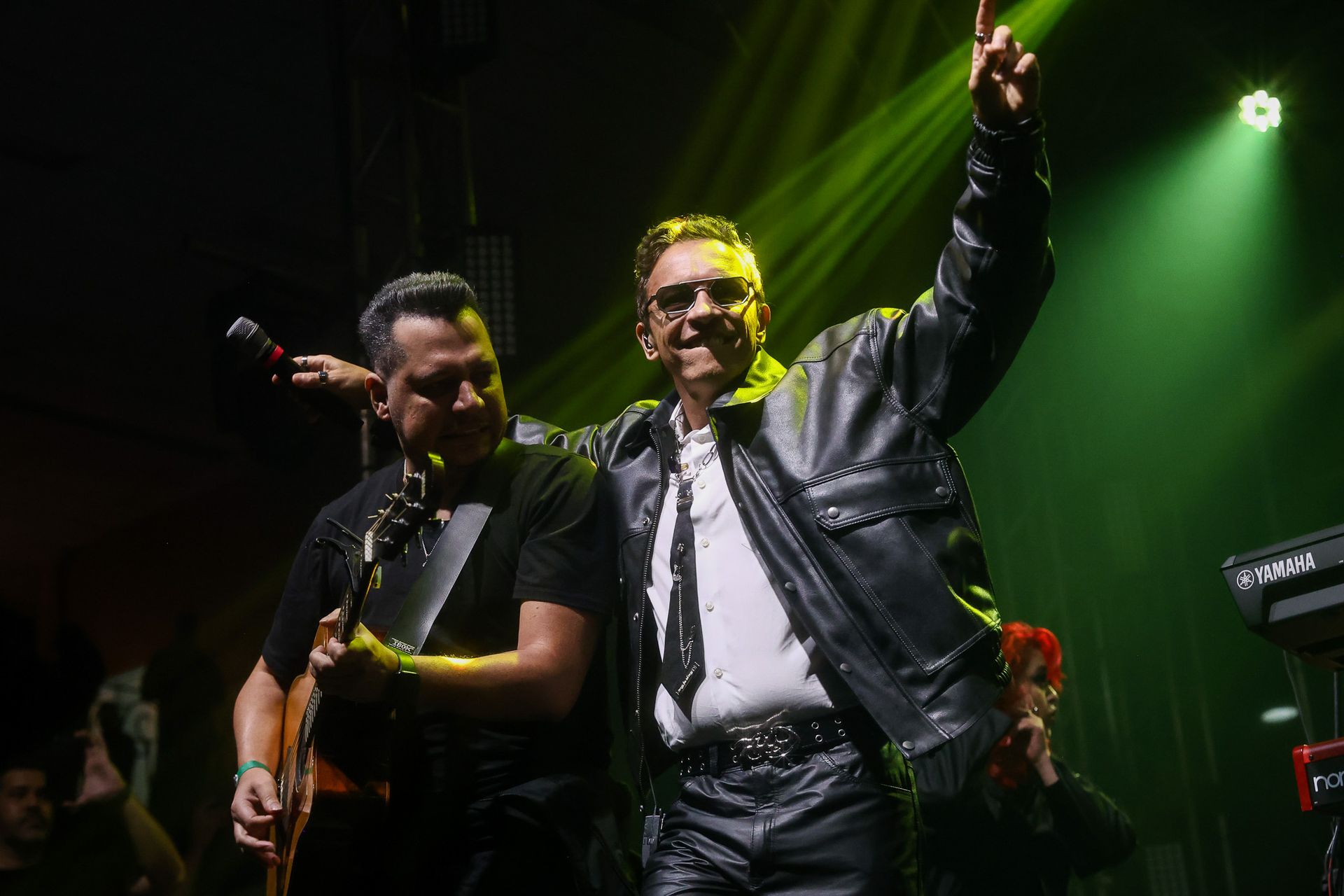 Dois músicos em um palco pouco iluminado; um toca guitarra enquanto o outro veste uma jaqueta de couro