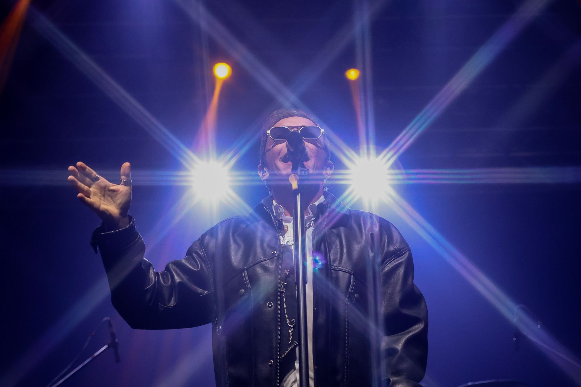 Um cantor de jaqueta escura está em pé diante de um microfone no palco, gesticulando com uma das mãos sob luzes brilhantes