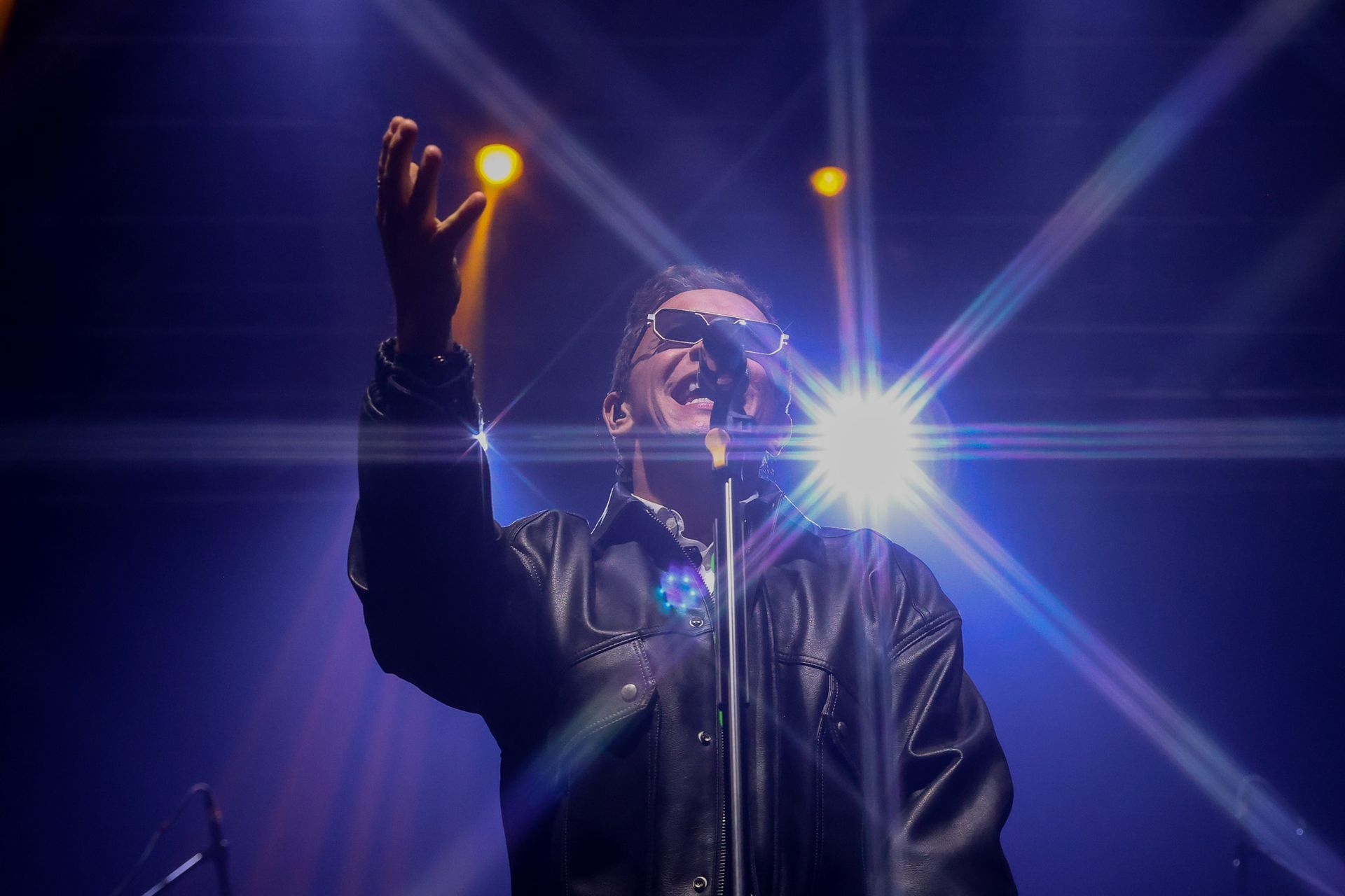 Um artista usando óculos escuros e jaqueta de couro se apresenta no palco, com um braço levantado, sob uma iluminação
