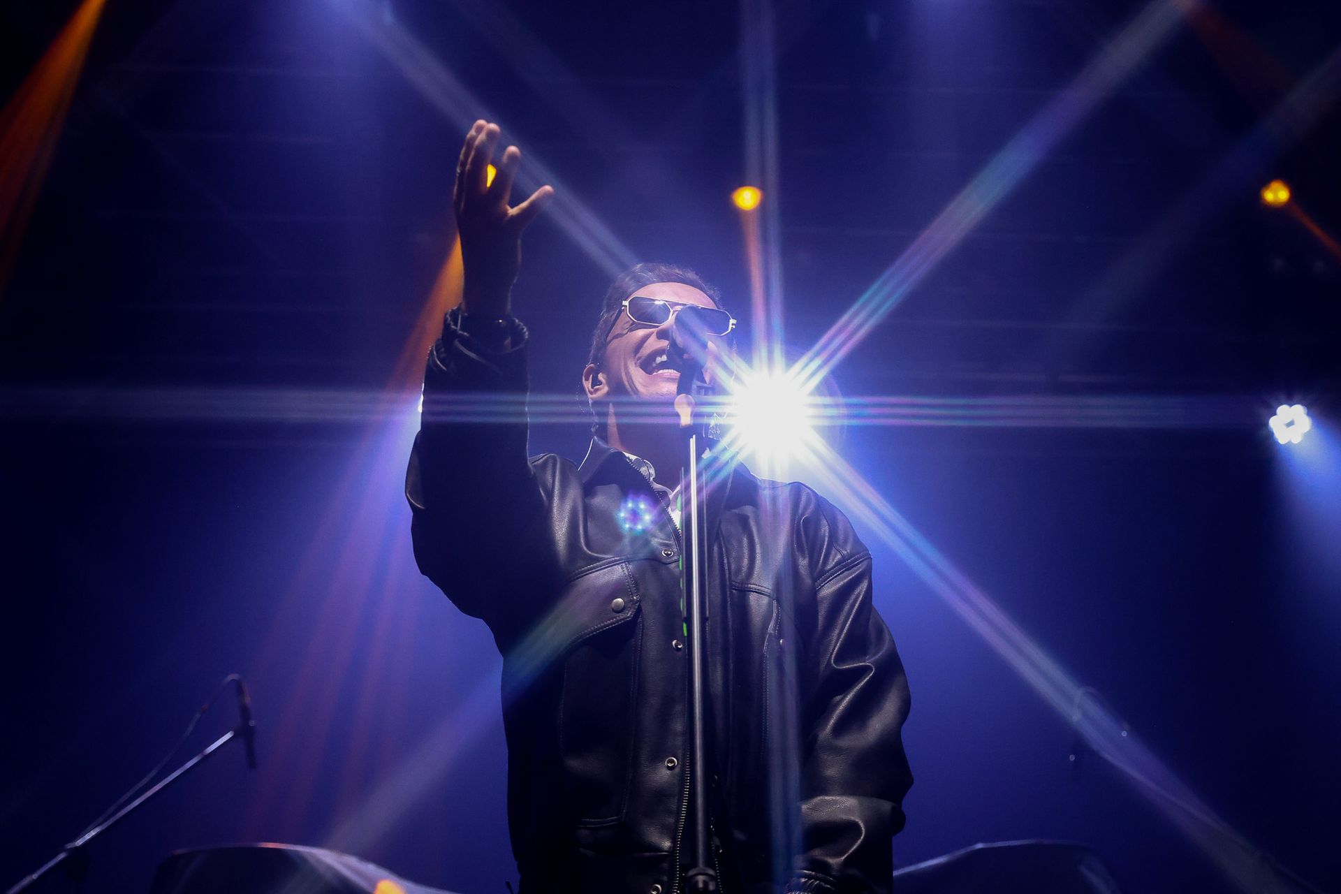 Um artista vestindo uma jaqueta de couro canta em um microfone no palco, iluminada por luzes de palco brilhantes.