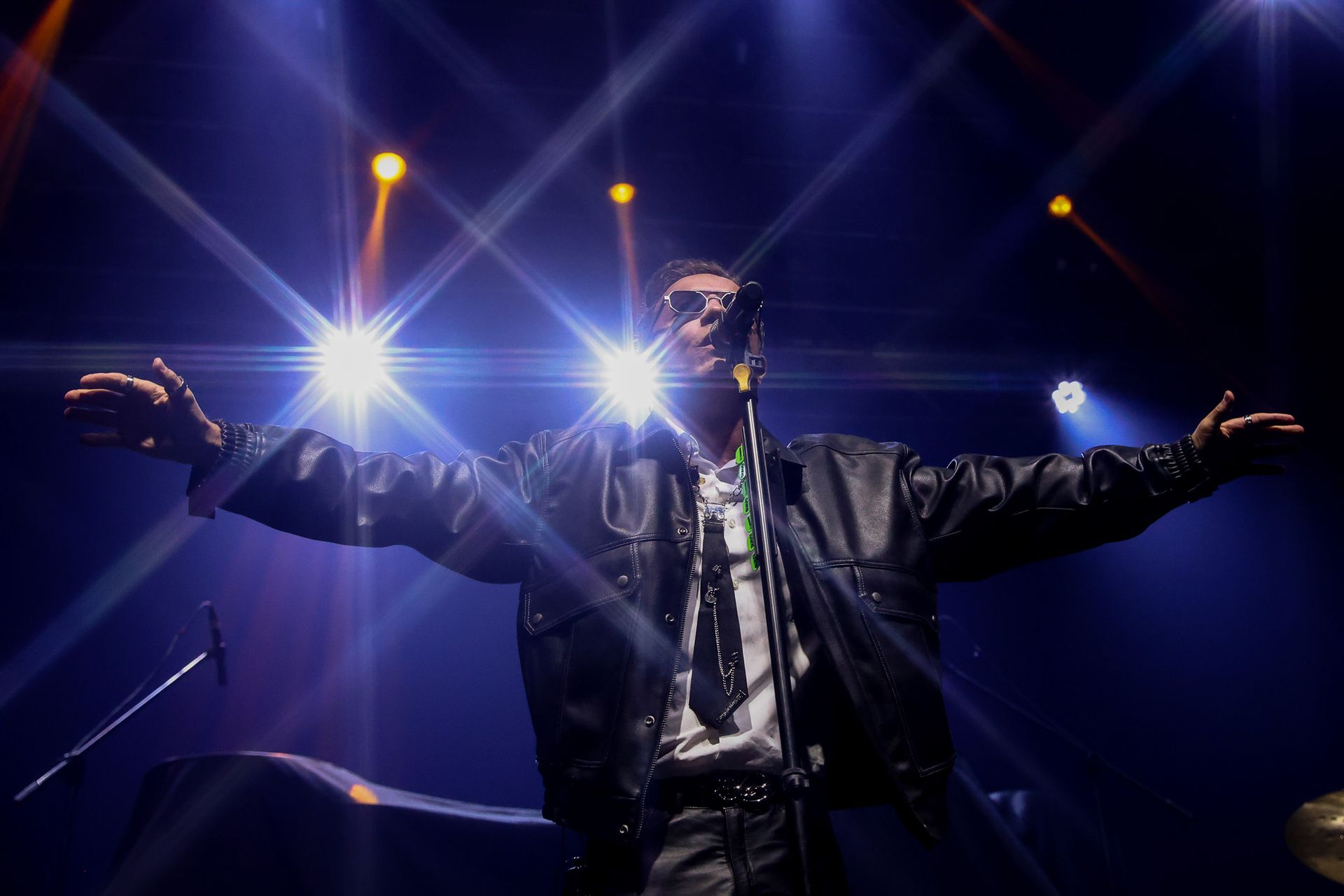Um cantor no palco, vestindo jaqueta de couro e gravata, com os braços estendidos em direção à plateia.