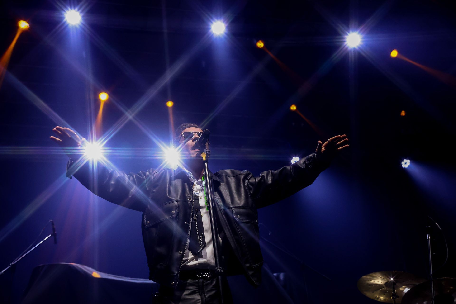 Um cantor vestindo uma jaqueta de couro escura se apresenta no palco com os braços estendidos sob holofotes brilhantes.