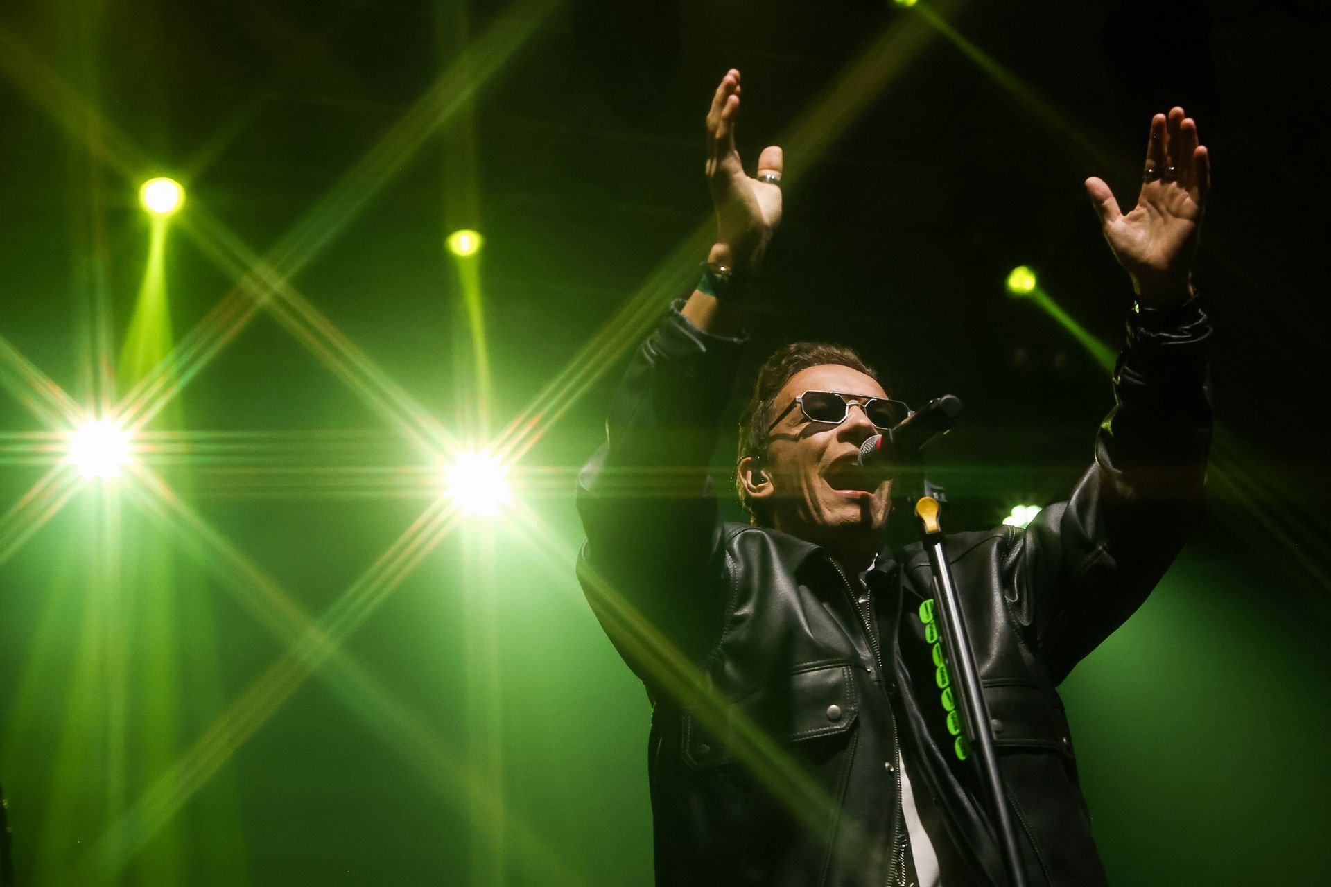Um cantor de jaqueta de couro se apresenta no palco com os braços erguidos, banhada por holofotes.