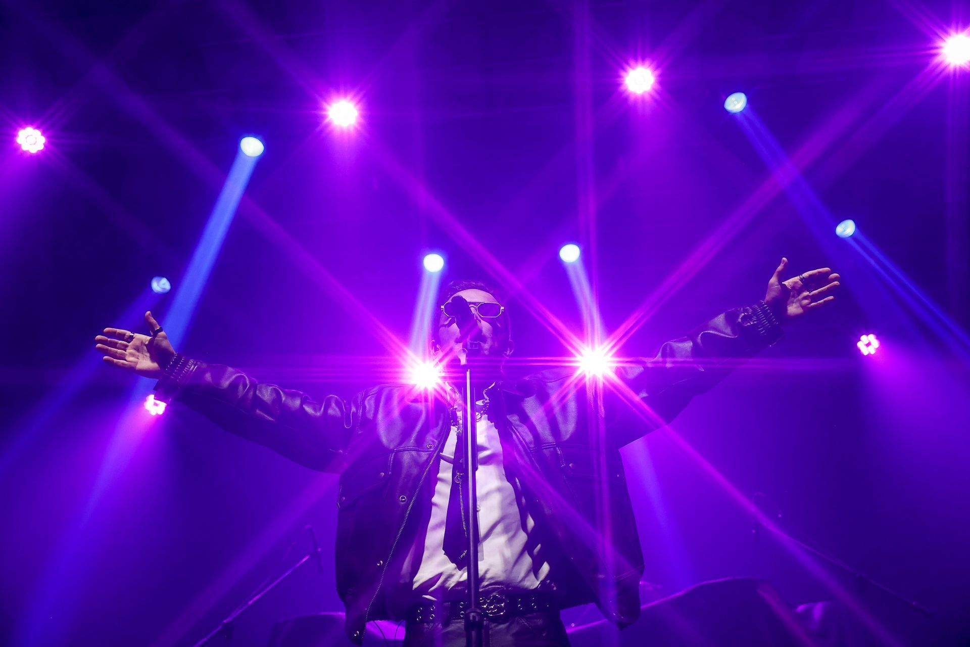 A silhueta de um artista com os braços estendidos em um palco, iluminada por holofotes roxos brilhantes e dramáticos.