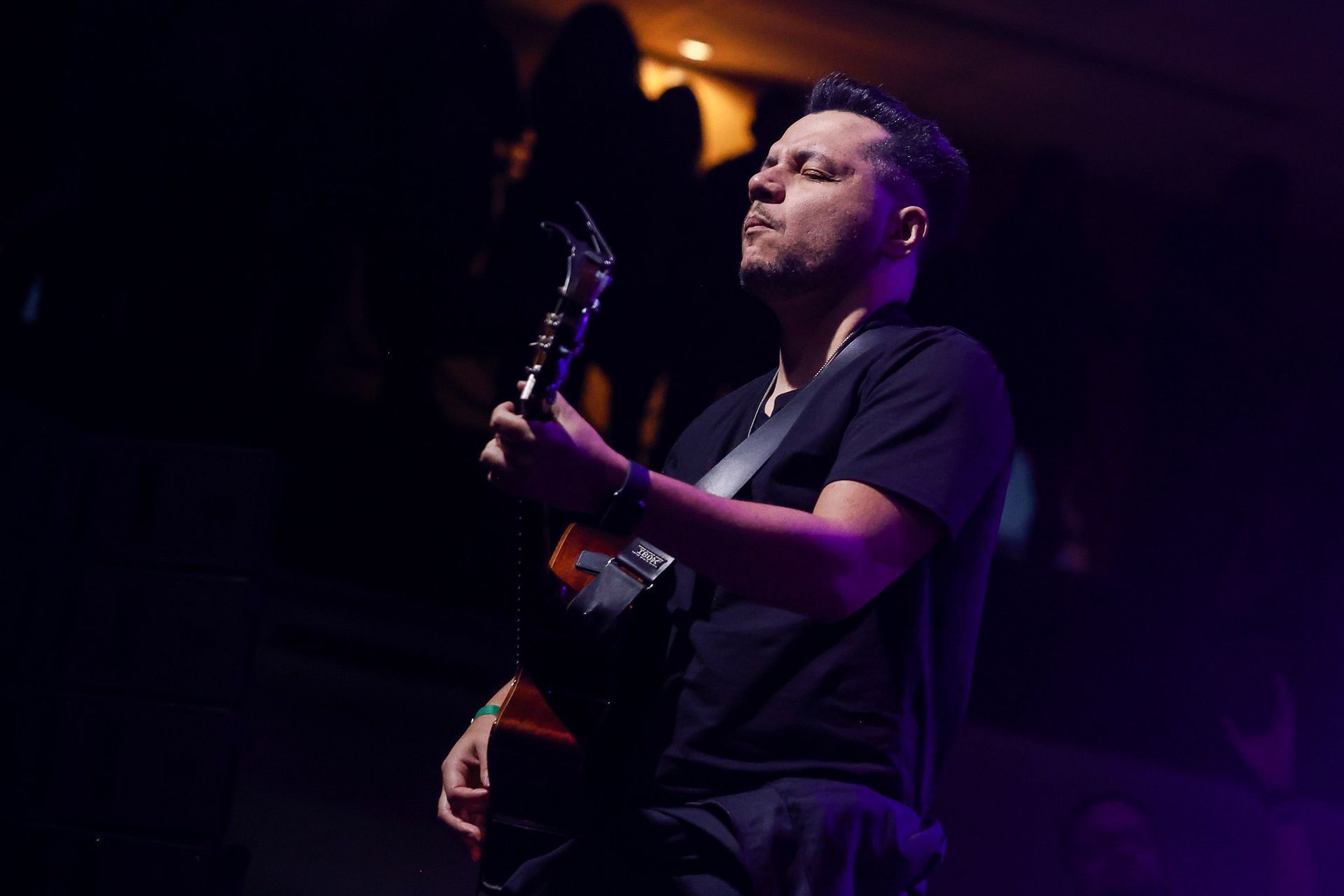 Um músico de olhos fechados toca violão em um palco com iluminação fraca e luz roxa.