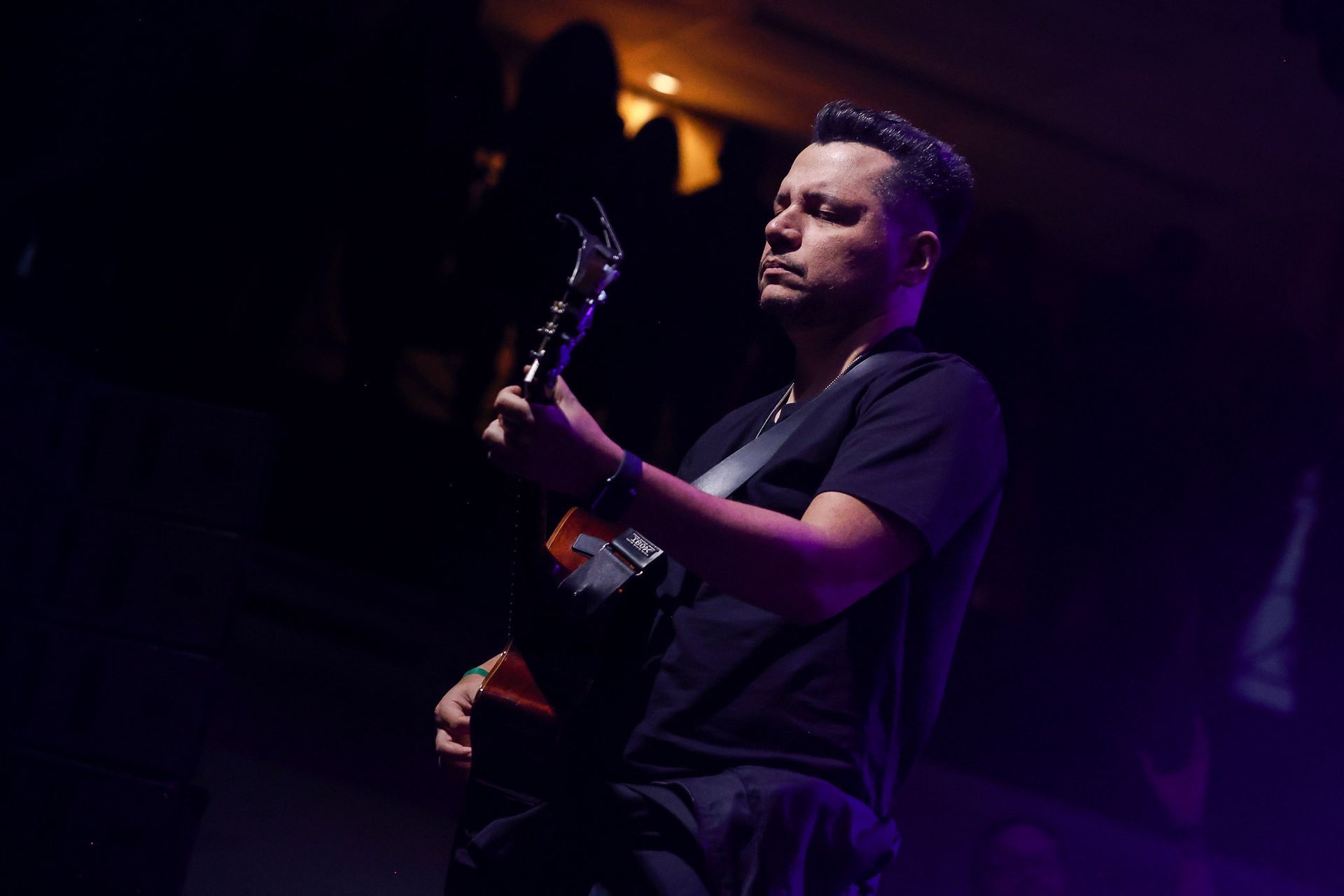 Um músico tocando violão em um palco escuro e pouco iluminado, com luzes de destaque na cor roxa.