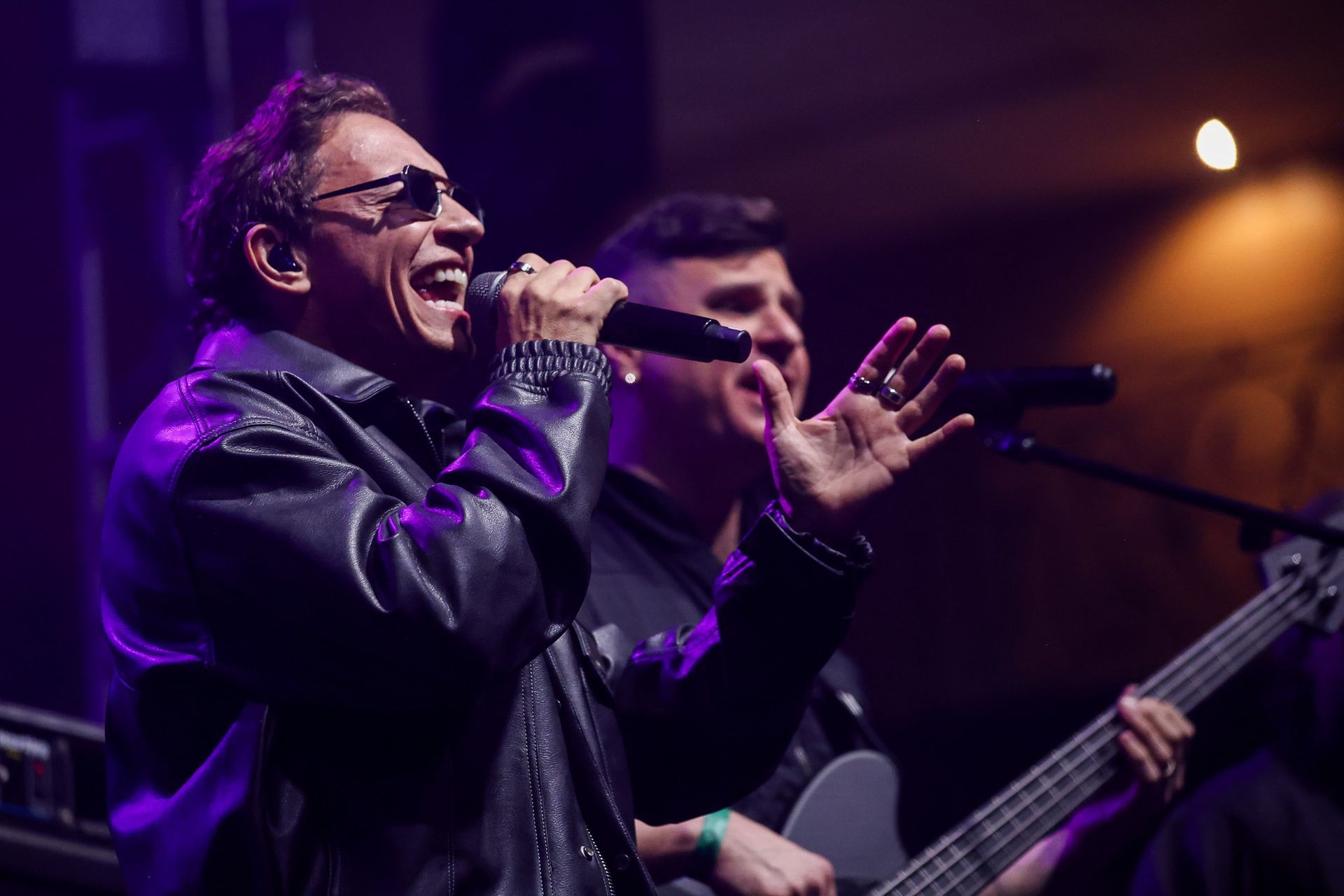 Um artista usando óculos escuros e jaqueta de couro canta em um microfone enquanto um membro da banda toca baixo no palco.