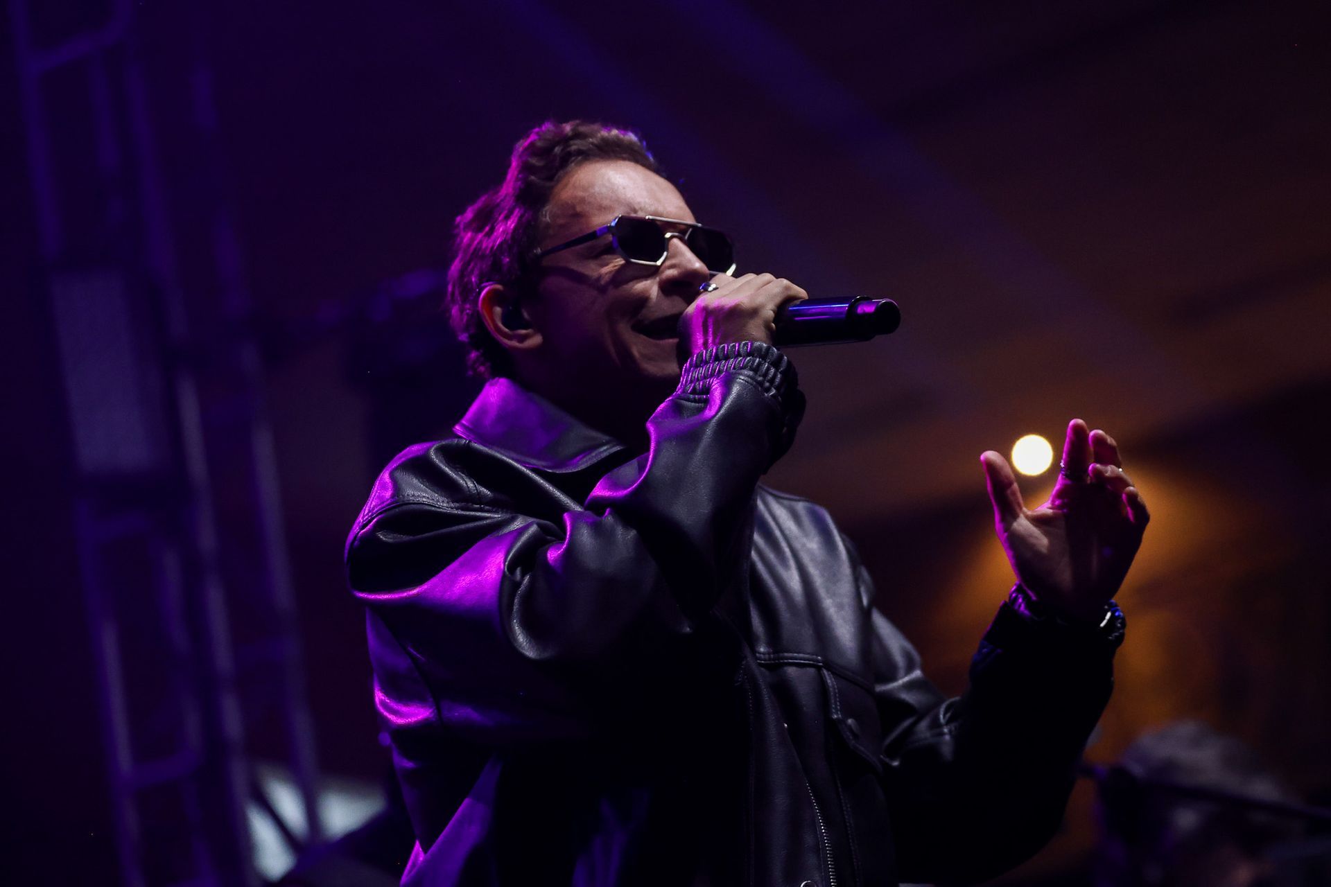 Um cantor de jaqueta de couro e óculos escuros se apresenta no palco,  e gesticulando com a mão direita.