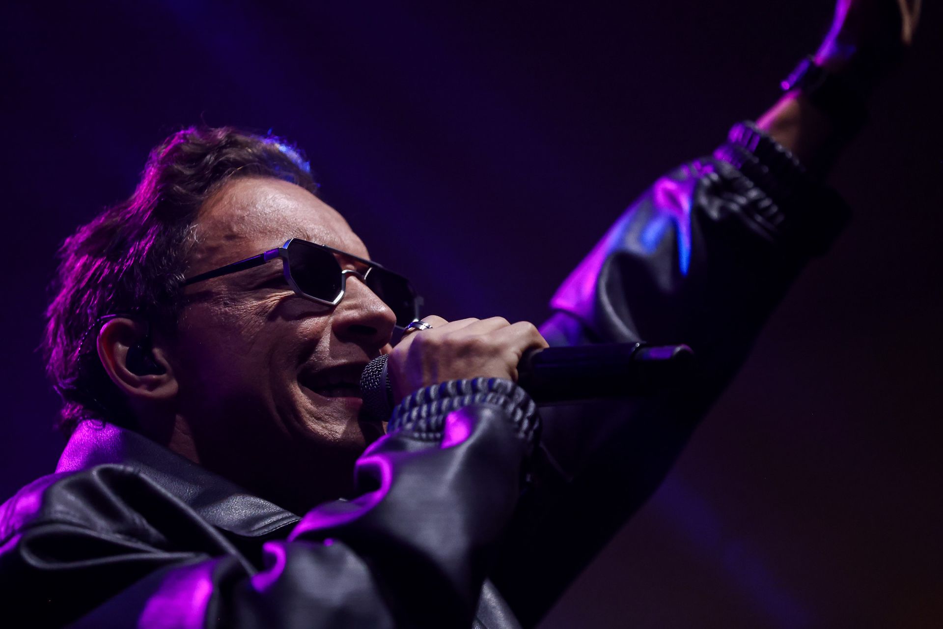 Um artista de óculos escuros e jaqueta de couro preta canta em um microfone com o braço levantado sob uma luz roxa no palco.