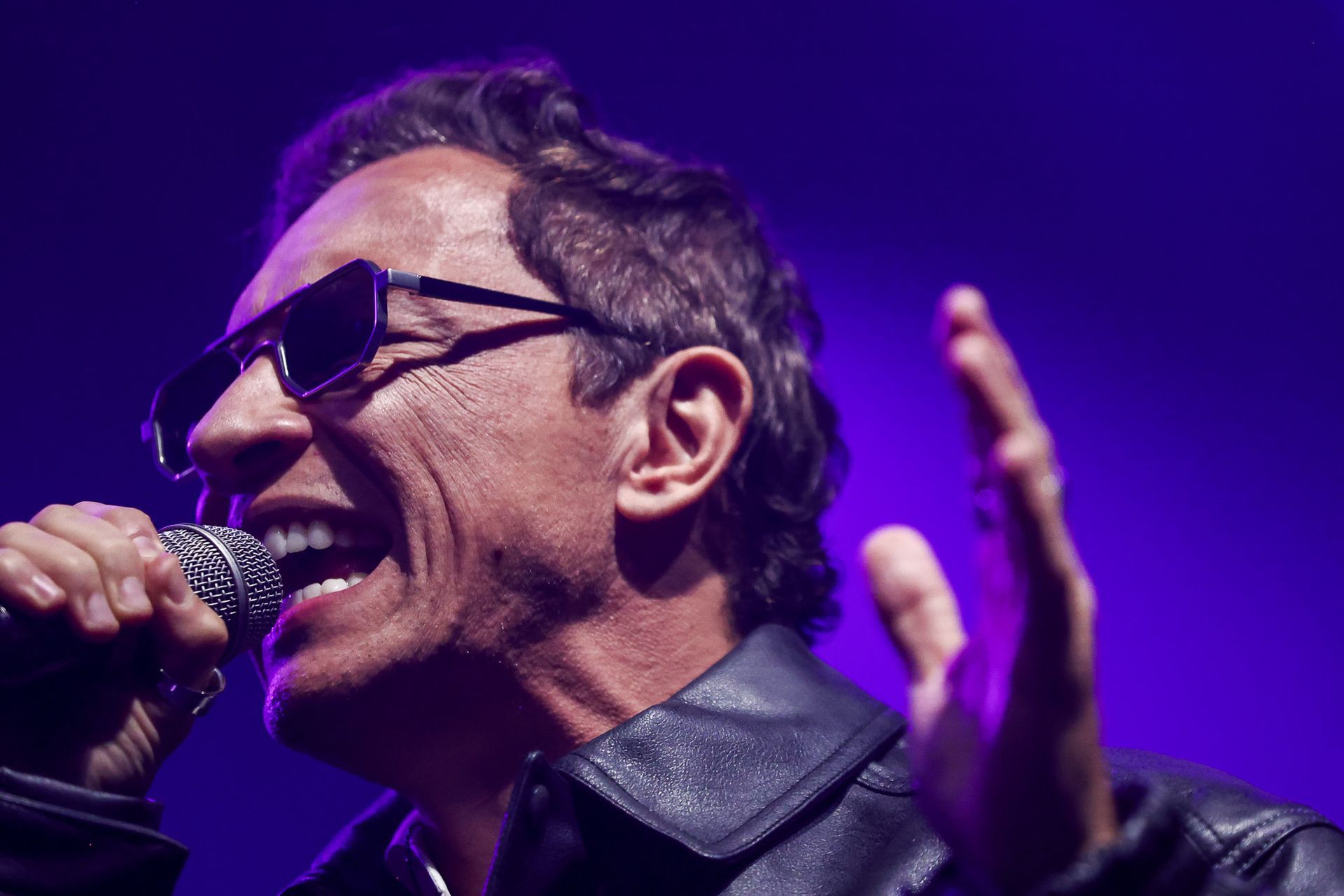 Um cantor de óculos escuros e jaqueta de couro se apresenta gesticulando com uma das mãos contra uma luz roxa.