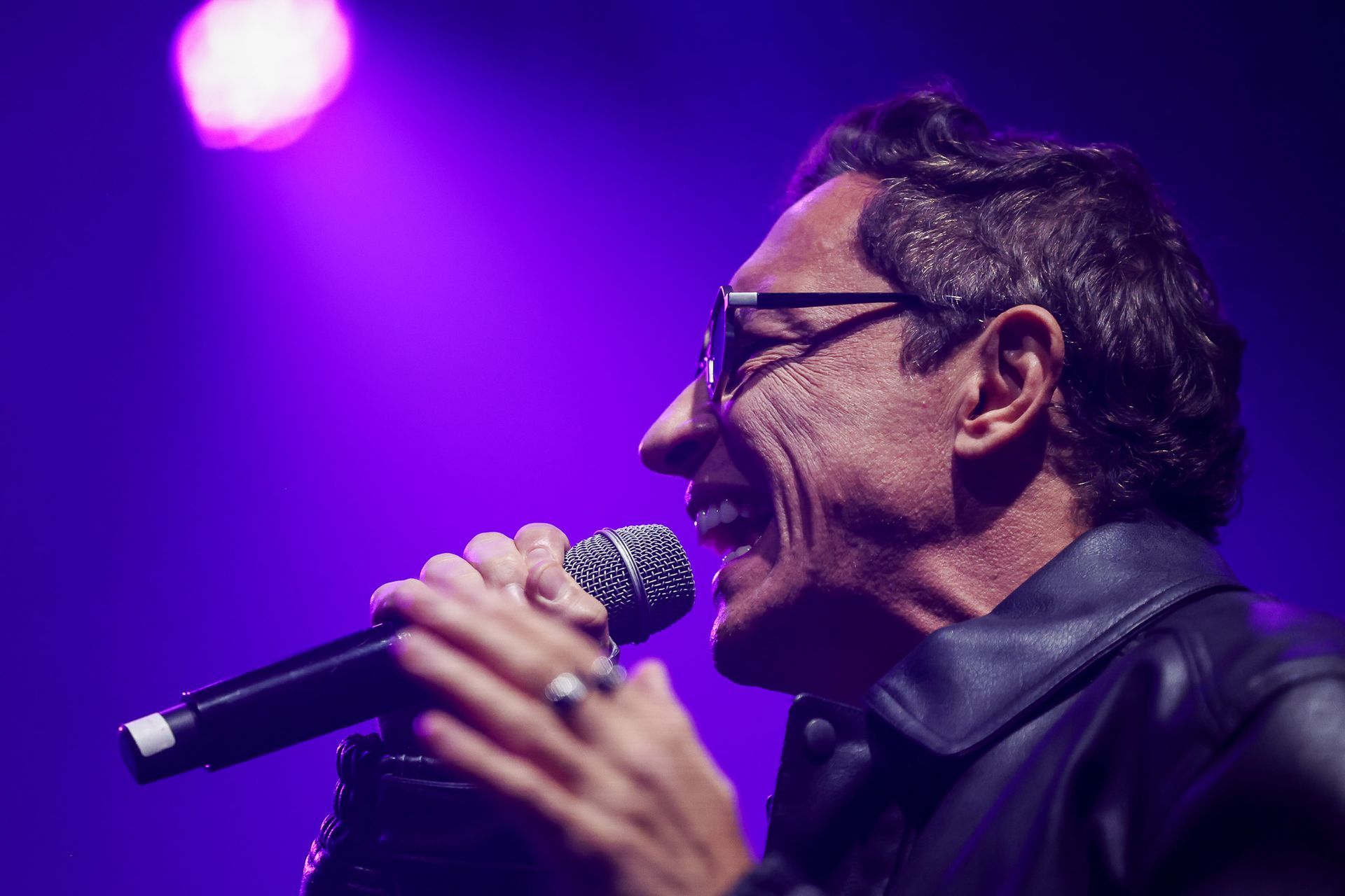 Um artista de óculos canta em um microfone sob luzes de palco roxas, sorrindo com os olhos fechados.