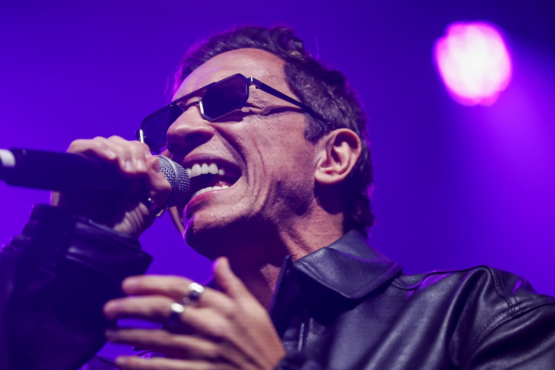 Um artista vestindo jaqueta de couro e óculos escuros cantando em um microfone em um palco iluminado com luz roxa.
