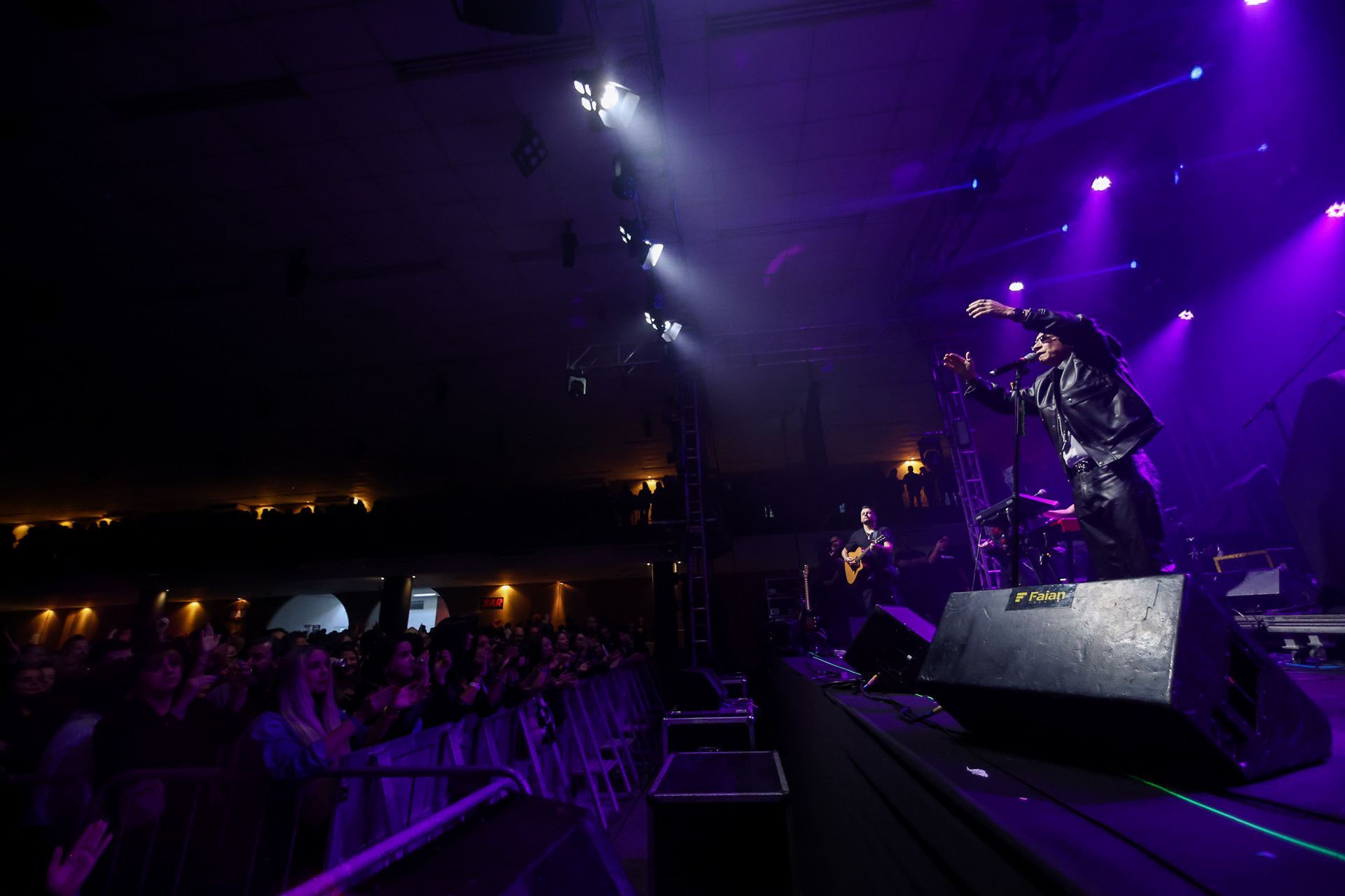 Um artista canta no palco durante um concerto, banhada por vibrantes luzes roxas, com uma plateia visível no local escuro.