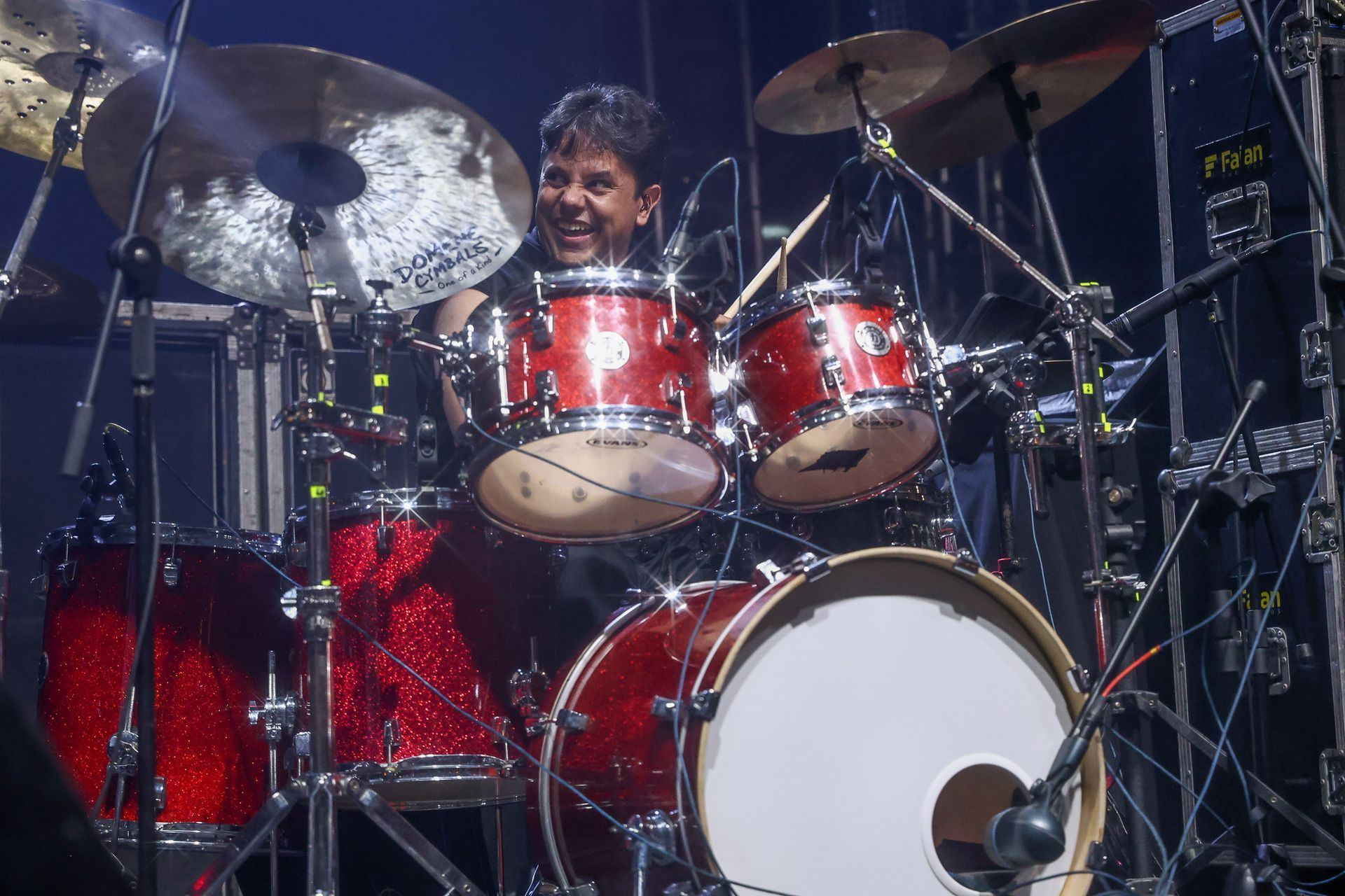 Um baterista sorridente toca uma bateria vermelha brilhante em um palco iluminado por luzes de concerto.